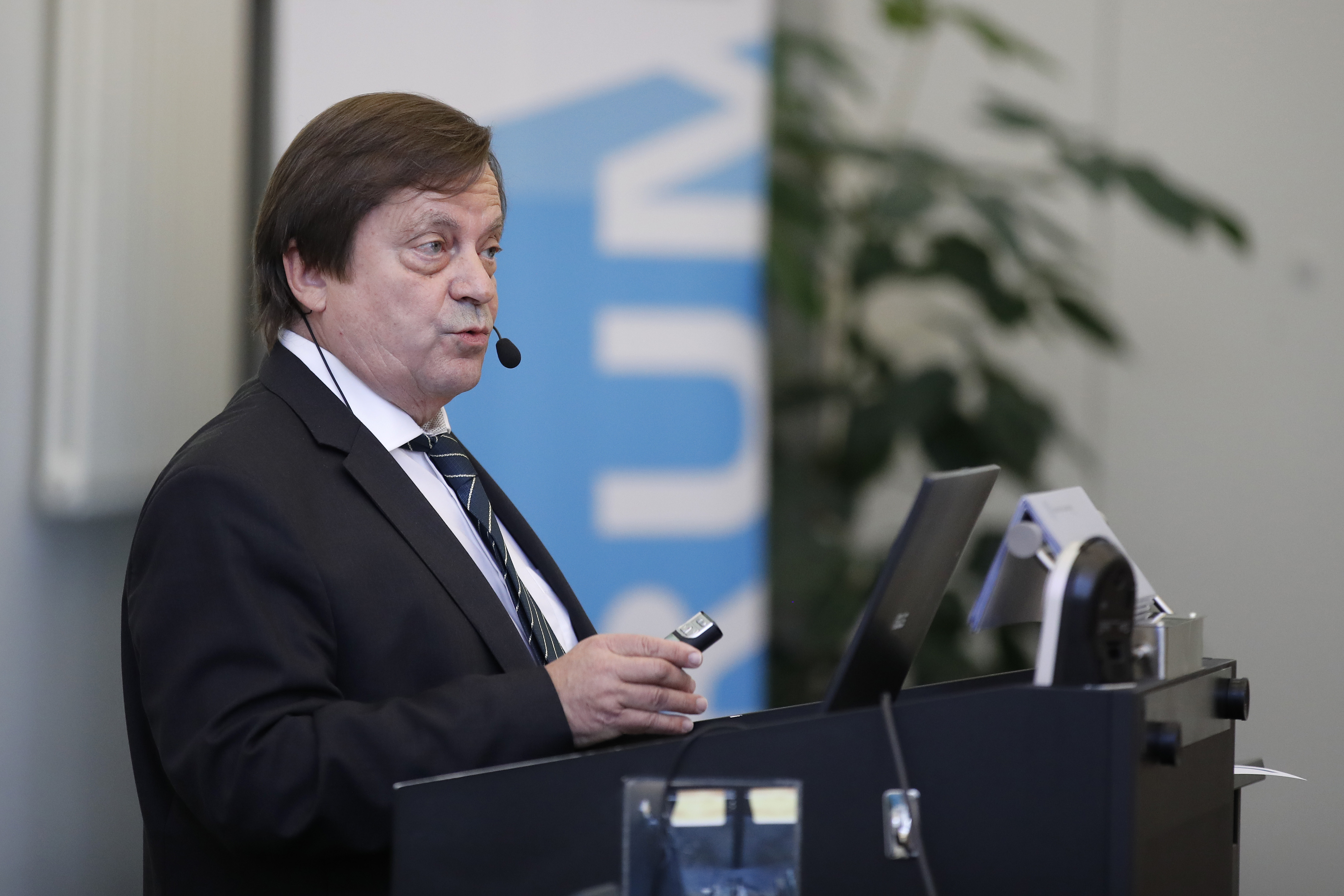 Dr. Andreas Zeller steht während seinem Referat am Rednerpult. Im Hintergrund ein blaues Plakat des  Forums für Universität und Gesellschaft