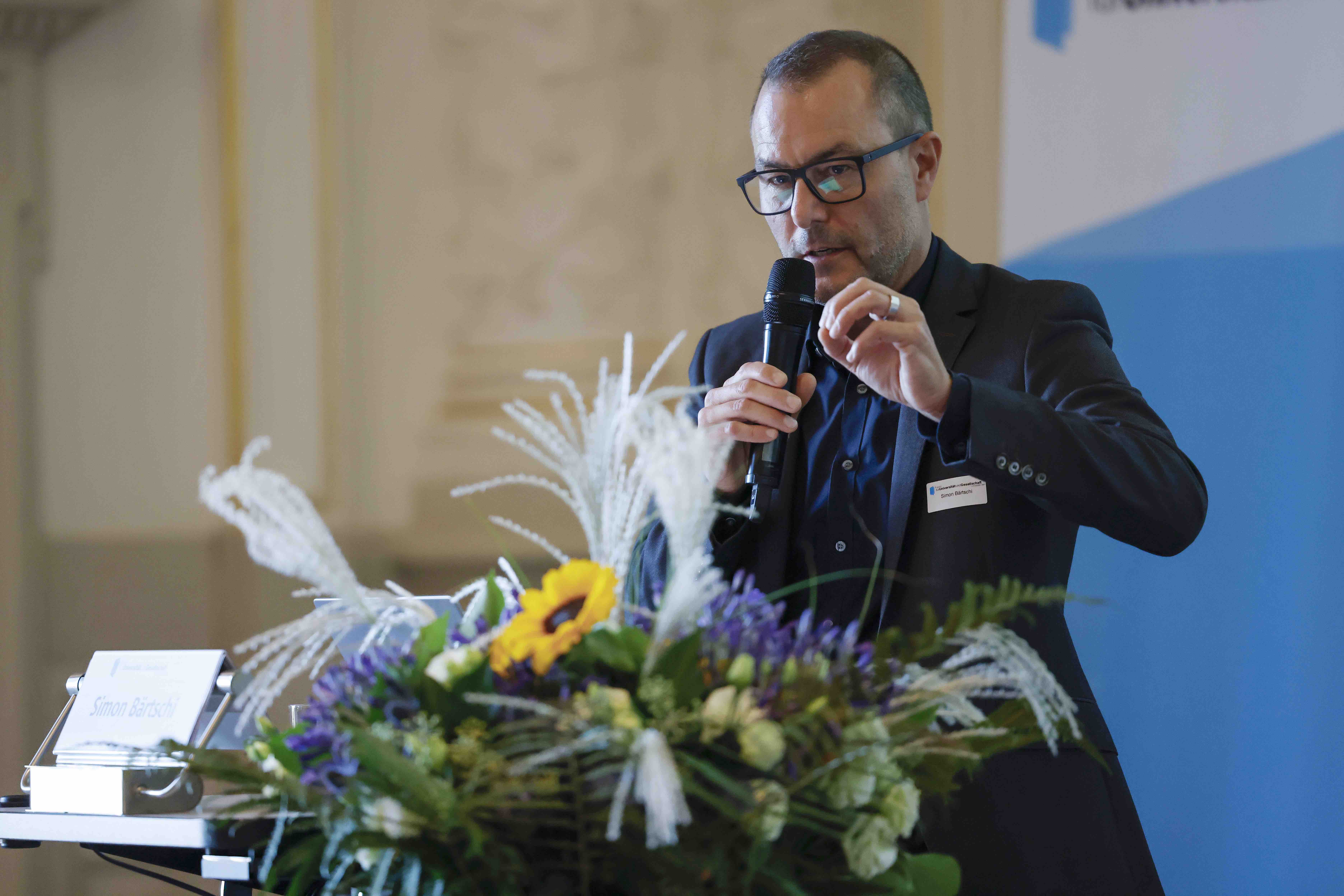 Der Referent Simon Bärtschi (Chefredaktor Berner Zeitung) steht während seinem Vortrag hinter dem Rednerpult, daneben ist ein Blumenstrauss abgestellt.