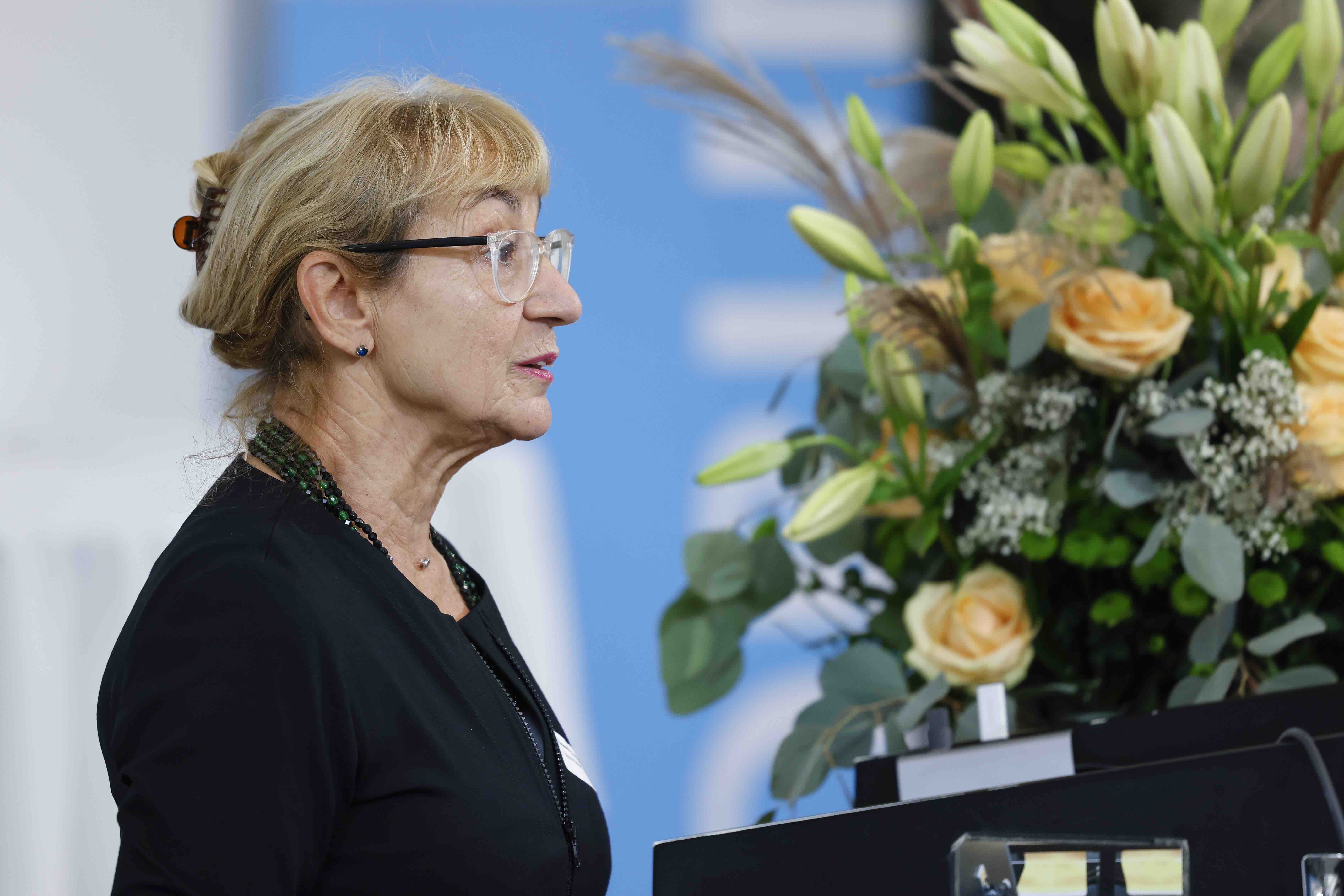 Seitliche Porträtaufnahme der Moderatorin Ursula Hürzeler. Sie trägt ein schwarzes Oberteil, im Hintergrund sind ein Blumenstrauss und ein Banner mit dem blauweissen Forumslogo sichtbar.