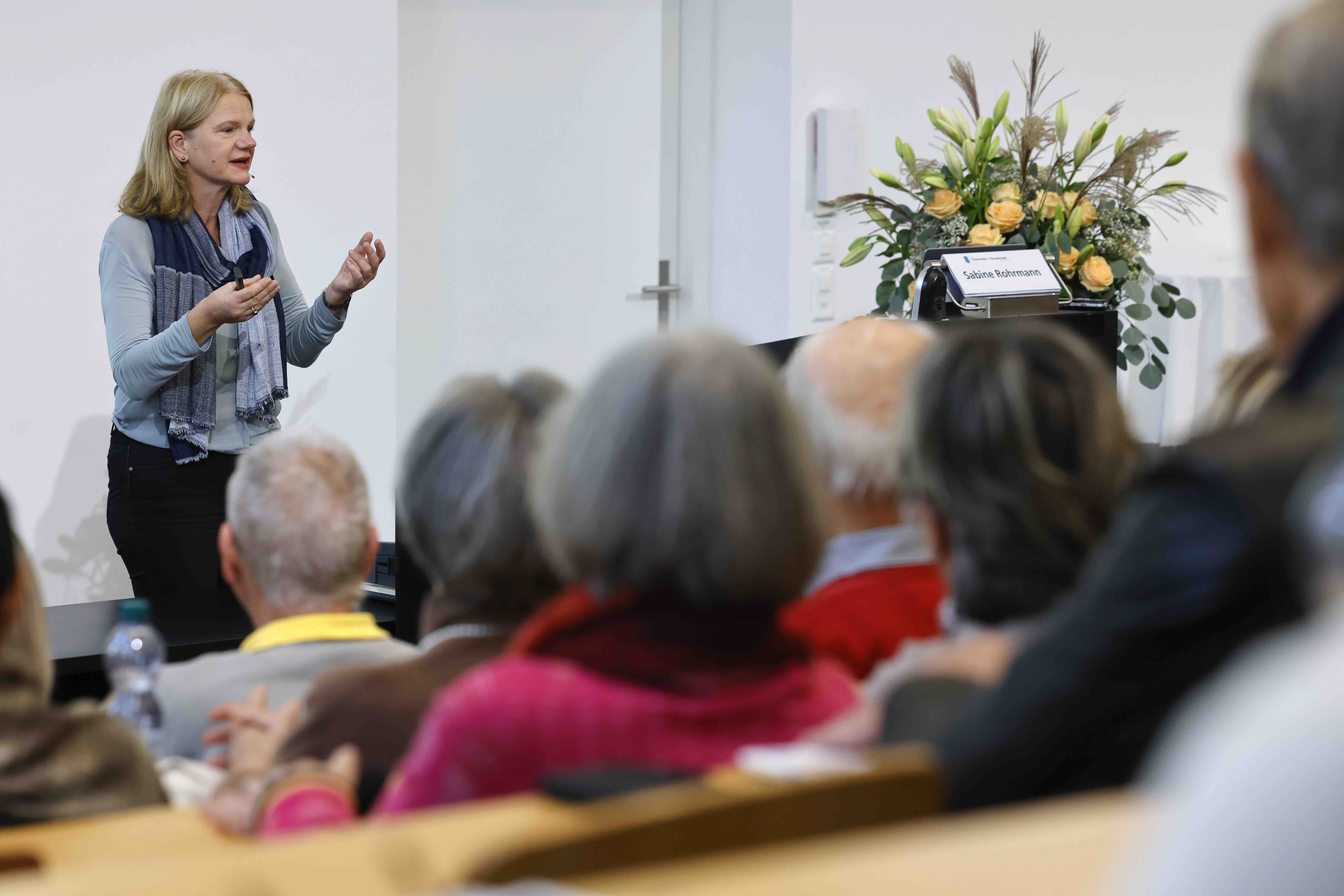 Links im Bild steht die Referentin Sabine Rohrmann und gestikuliert mit beiden Händen. Rechts im Bild ist ein Blumenstrauss und im Vordergrund ein Teil des Publikums zu sehen.