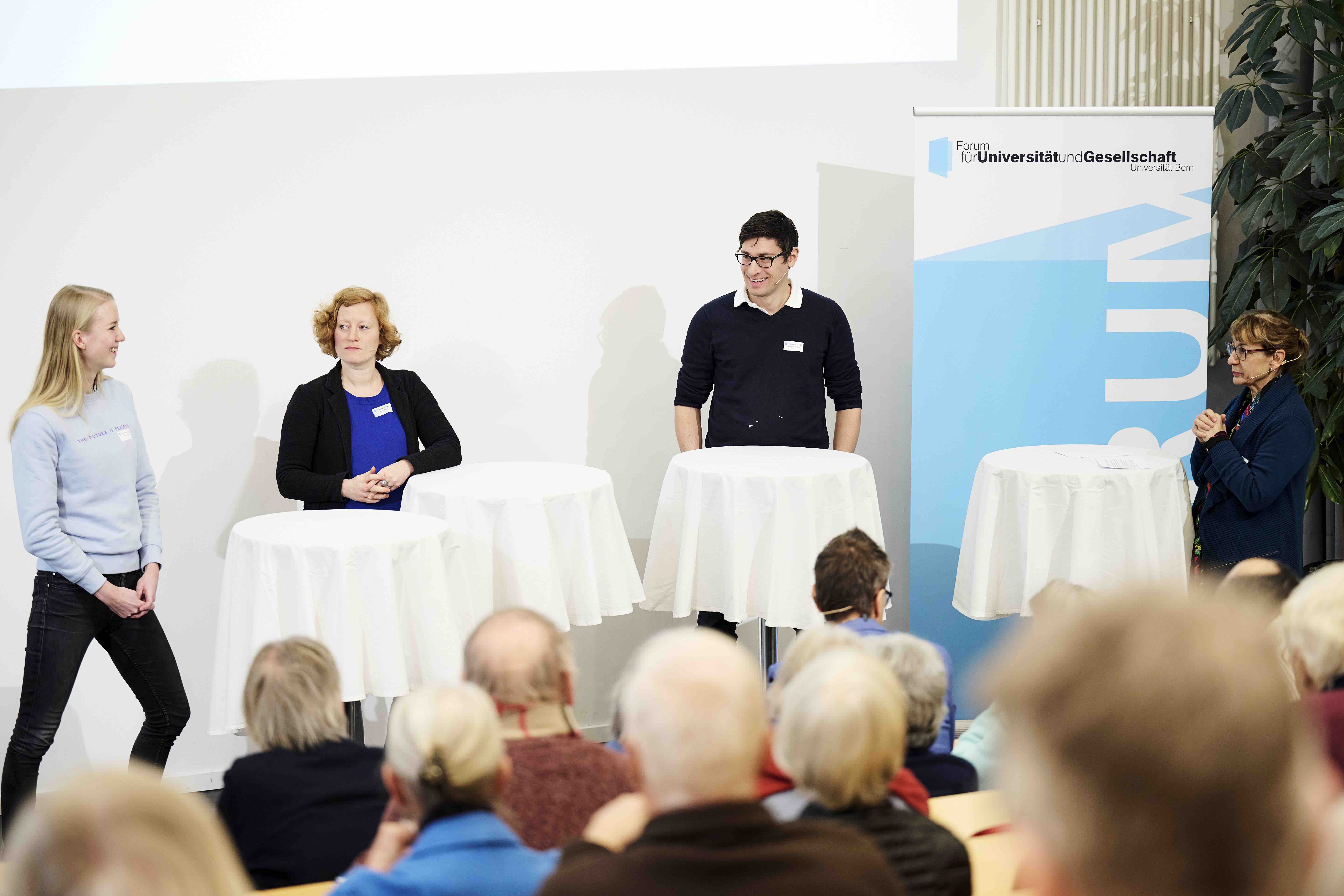 Zwei Frauen und ein Mann stehen während einer Podiumsdiskussion hinter Stehtischen mit weissen Tischtüchern. Im Hintergrund ist ein Banner mit dem blauweissen Forumslogo sichtbar.