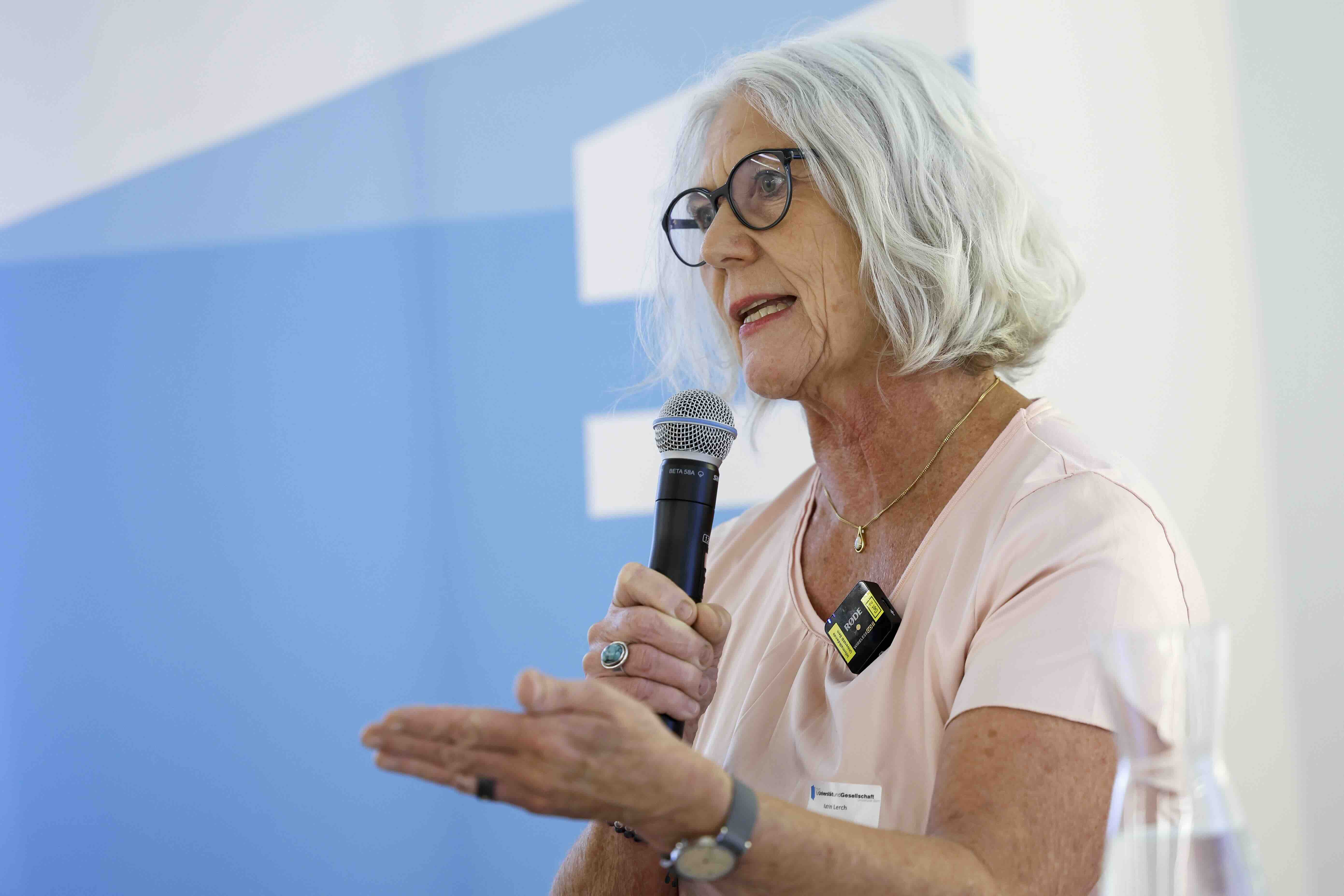 Eine Frau mit grauen, schulterlangen Haaren und Brille spricht in ein Mikrofon. Sie trägt eine helle Bluse, eine Halskette und eine Armbanduhr. Mit der freien Hand gestikuliert sie. Im Hintergrund ist ein blau-weißes Banner des Forums für Universität und Gesellschaft sichtbar.