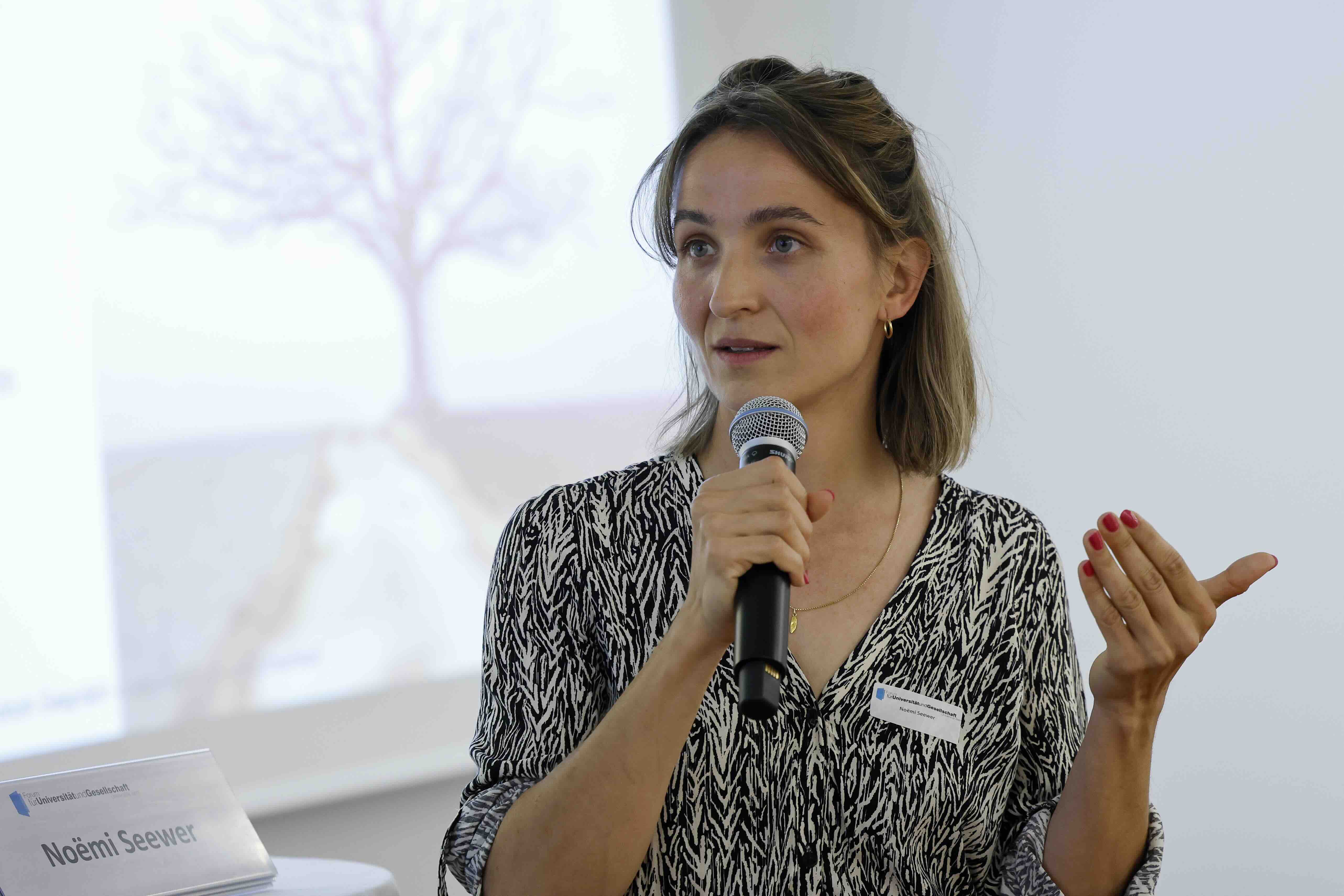 Eine Frau mit schulterlangen, braunen Haaren spricht in ein Mikrofon. Sie trägt eine schwarz-weiß gemusterte Bluse und eine dezente Halskette. Mit der freien Hand gestikuliert sie. Im Hintergrund ist unscharf eine Projektion mit dem Bild eines Baumes zu sehen
