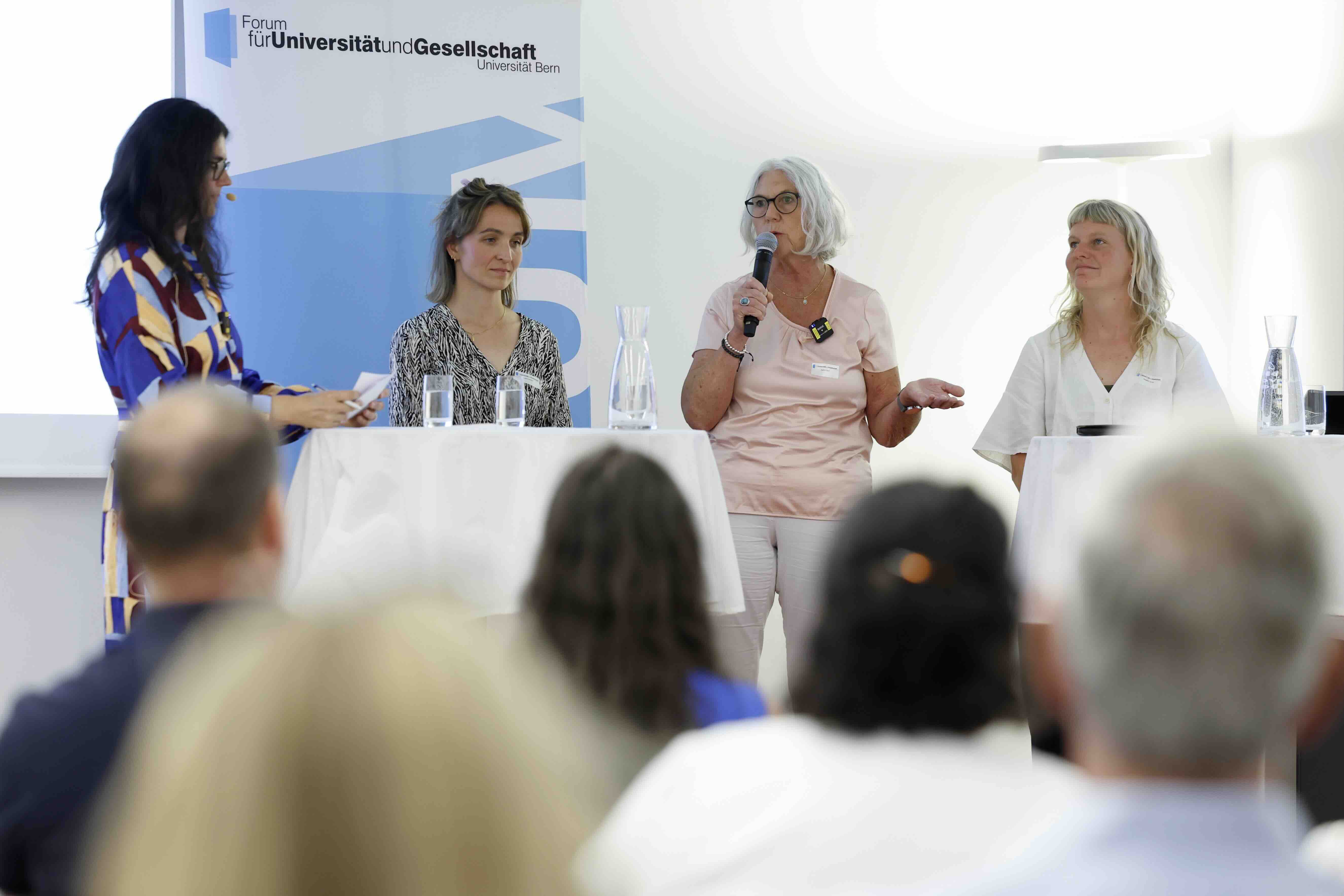 Auf dem Foto sind vier Frauen bei einer Podiumsdiskussion zu sehen. Sie stehen bzw. sitzen hinter zwei runden, mit weißen Tischdecken bedeckten Stehtischen. uf den Tischen stehen Glaskaraffen und Gläser mit Wasser. Im Hintergrund ist ein Roll-up mit dem Logo des „Forum für Universität und Gesellschaft Universität Bern“ sichtbar. Im Vordergrund sind unscharf die Köpfe des Publikums zu erkennen.