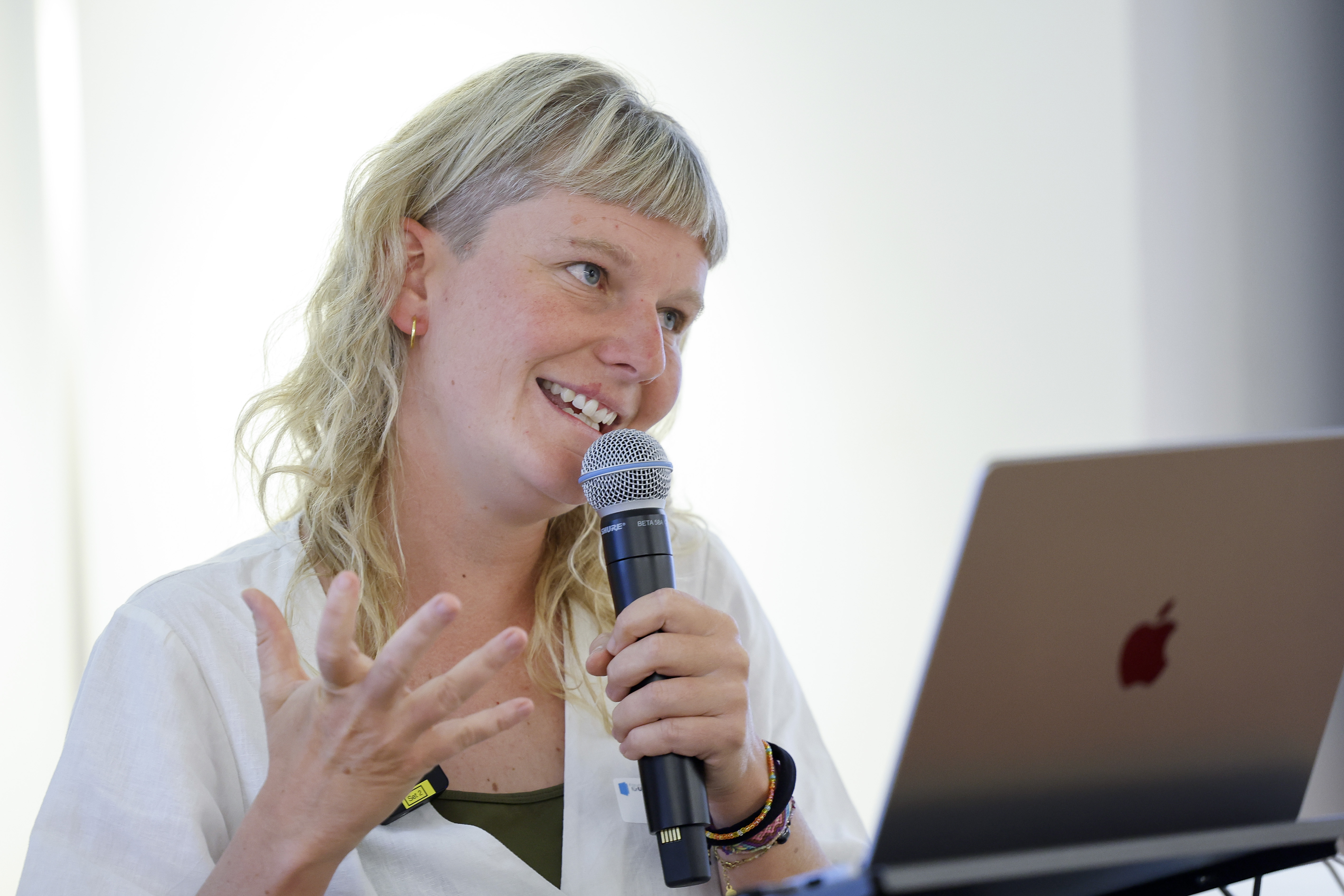 Eine Frau mit hellblonden, schulterlangen Haaren spricht lächelnd in ein Mikrofon. Sie trägt eine weiße Bluse und gestikuliert mit einer Hand. Vor ihr steht ein aufgeklappter Laptop.