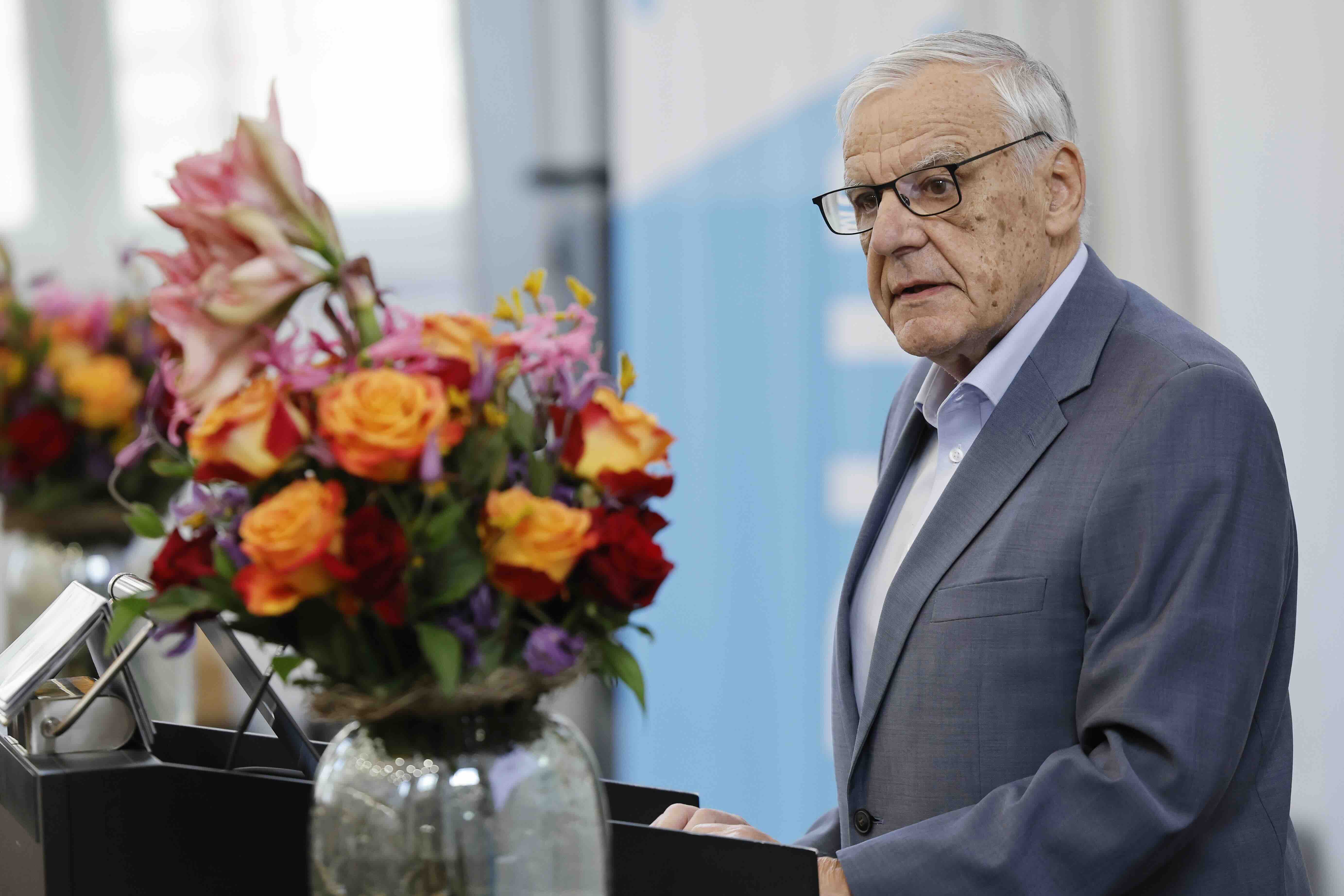 Ein älterer Mann mit grauen Haaren und Brille steht an einem Rednerpult und spricht. Er trägt einen grauen Anzug und schaut aufmerksam in den Raum. Neben ihm steht ein großer Blumenstrauß mit orangefarbenen, rosa und roten Blüten in einer Glasvase. Der Hintergrund ist hell und unscharf.