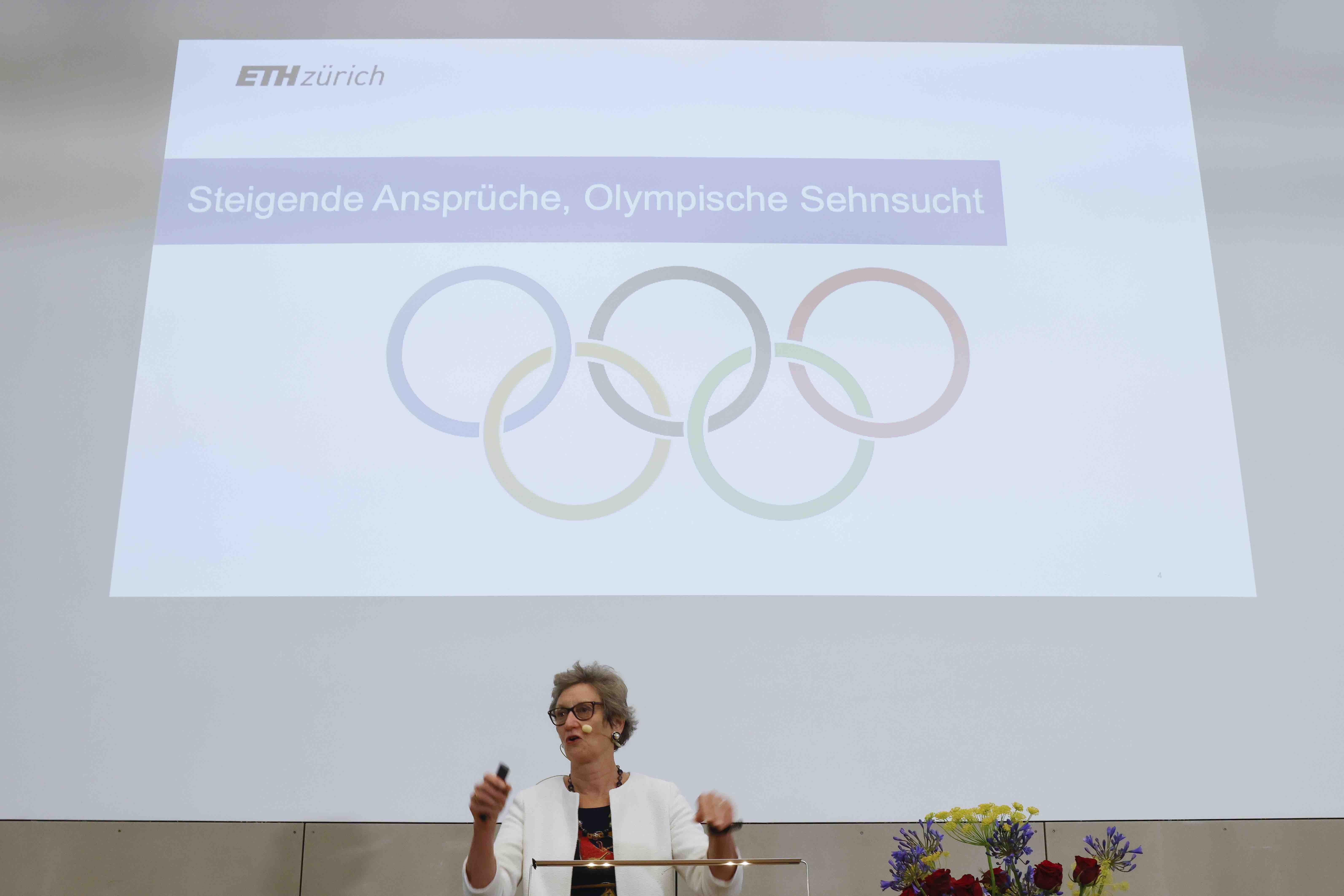 Professorin Sarah Springman während ihrem Referat in Bern. Hinter ihr ist die Projektion ihrer PowerPoint-Präsentation sichtbar. Sie zeigt die fünf Olympischen Ringe und den Titel «Steigende Ansprüche, Olympische Sehnsucht».