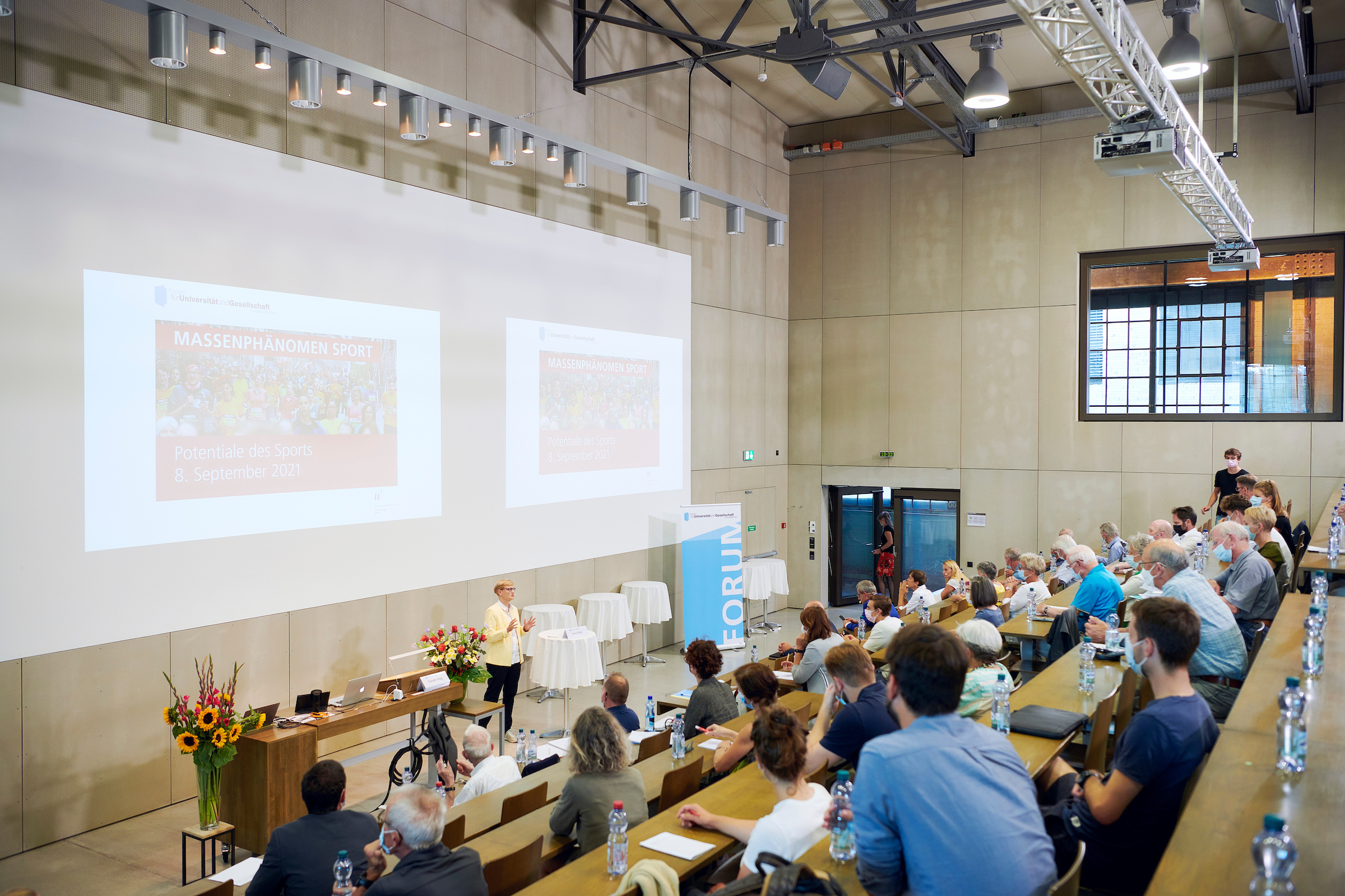 Das Publikum sitzt im Hörsaal der Universität Bern und lauscht der Begrüssung der Moderatorin Susy Schär, die vorne am Rednerpult steht