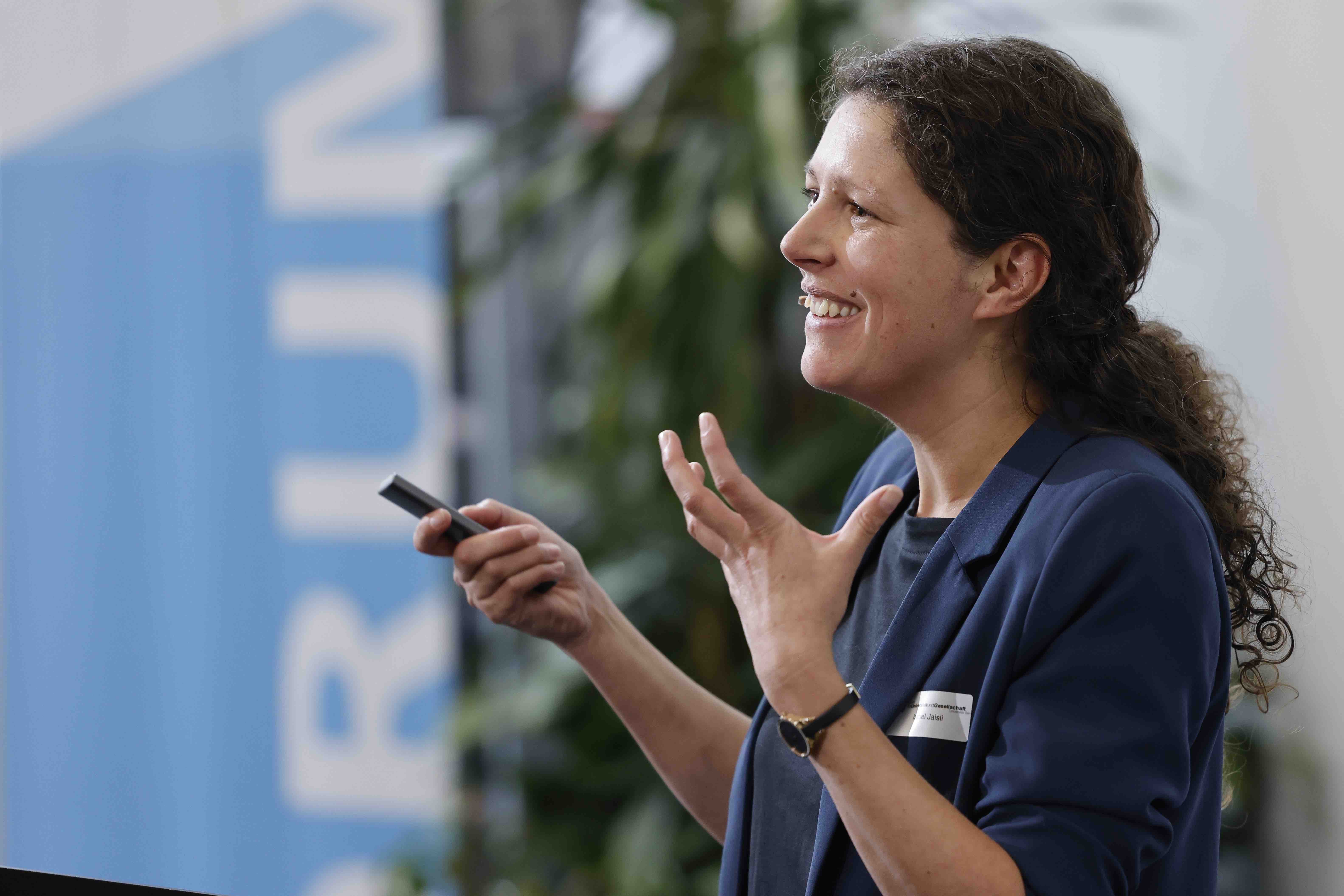 Porträtaufnahme von Isabel Jaisli während ihrem Referat. Sie spricht und gestikuliert; im Hintergrund sind eine grüne Pflanze und ein Banner mit dem blauweissen Forumslogo zu sehen.