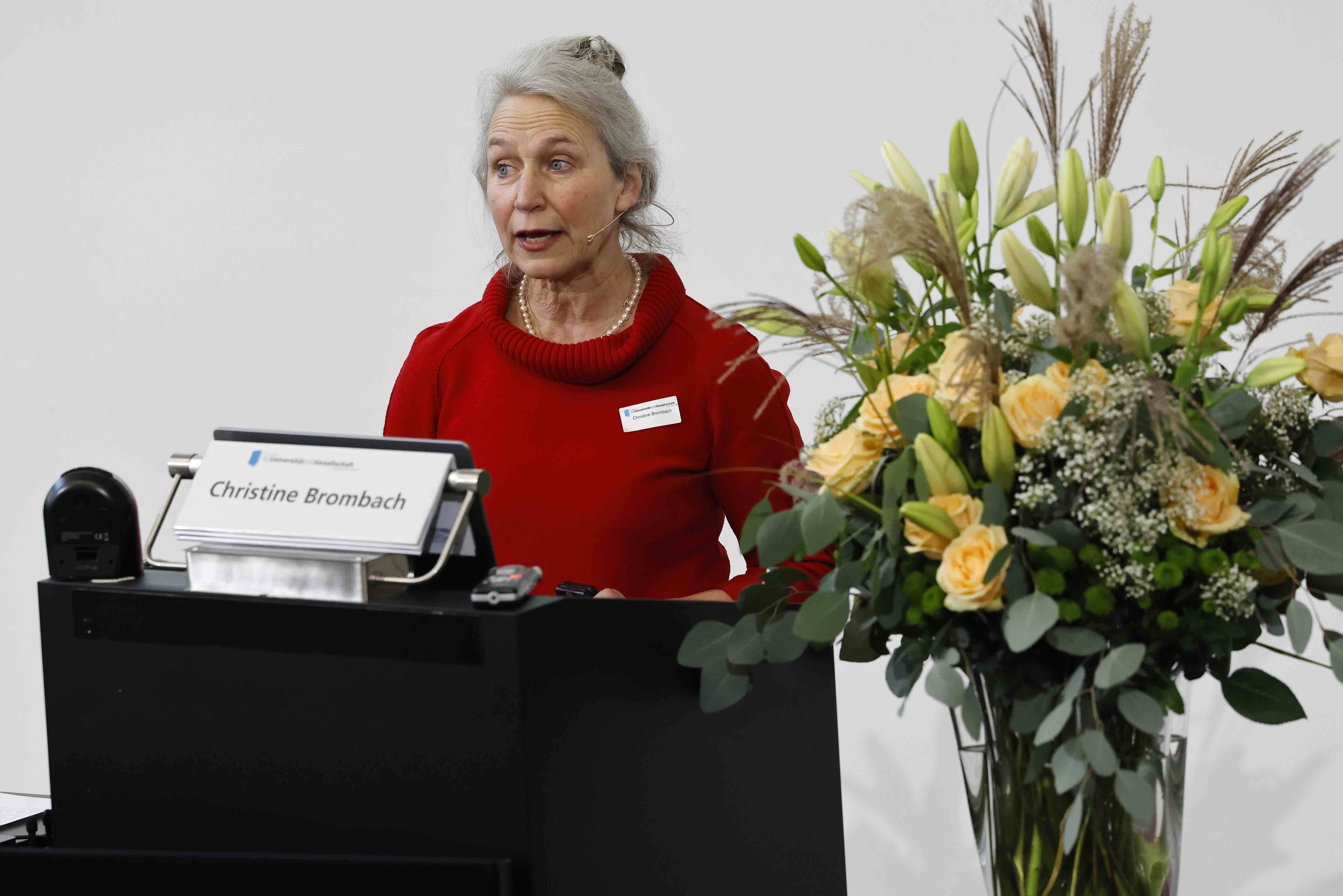 Aufnahme der Referentin Prof. Dr. Christine Brombach hinter einem Rednerpult. Sie trägt ein rotes Kleid, neben ihr ist ein Blumenstrauss in beige- und Gelbtönen zu sehen.
