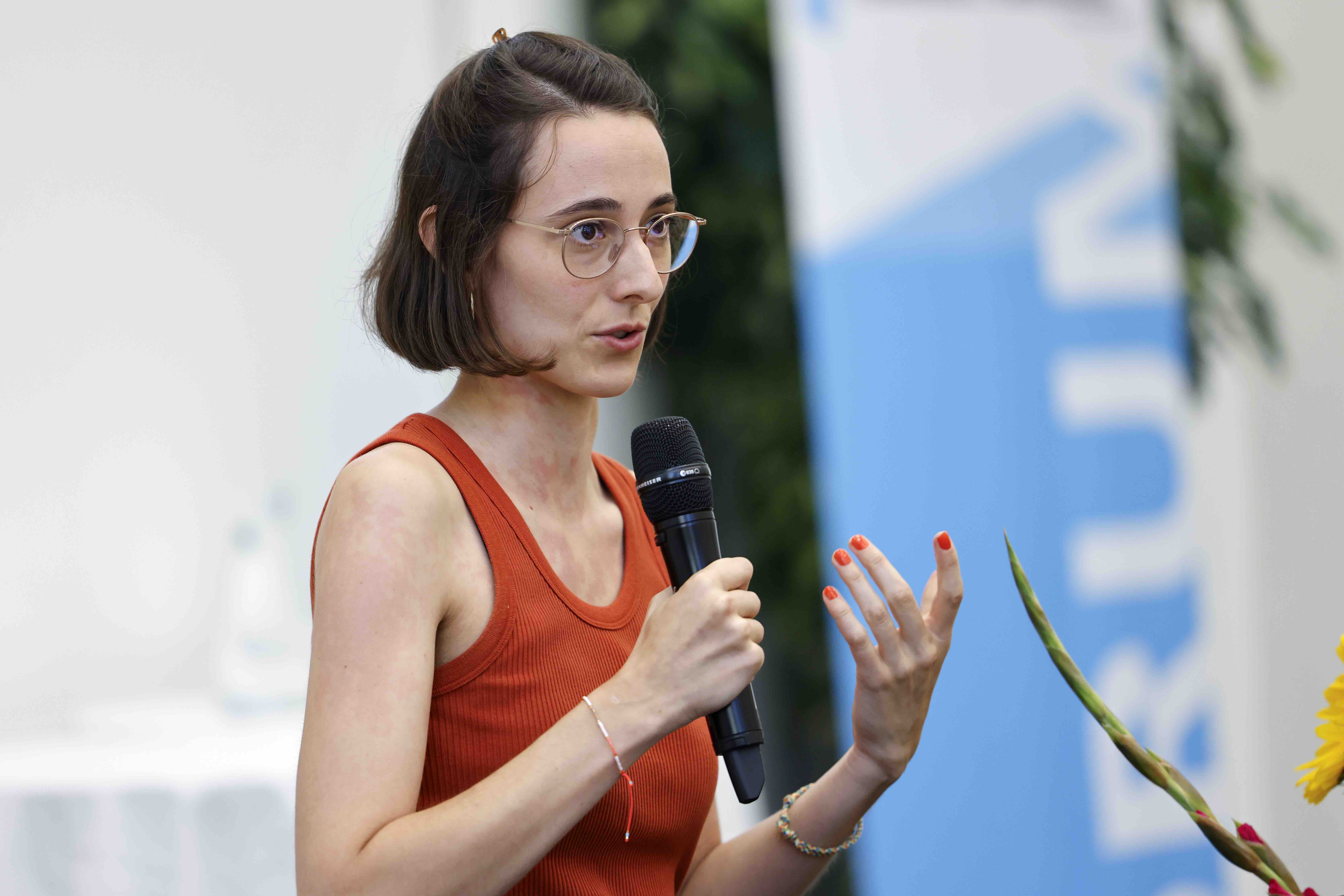 Die Referentin Janine Bürkli spricht in ein Handmikrofon und lacht. Im Hintergrund sind ein Blumenstrauss, grüne Zimmerpflanzen und ein blauweisses Banner mit dem Forumslogo erkennbar.