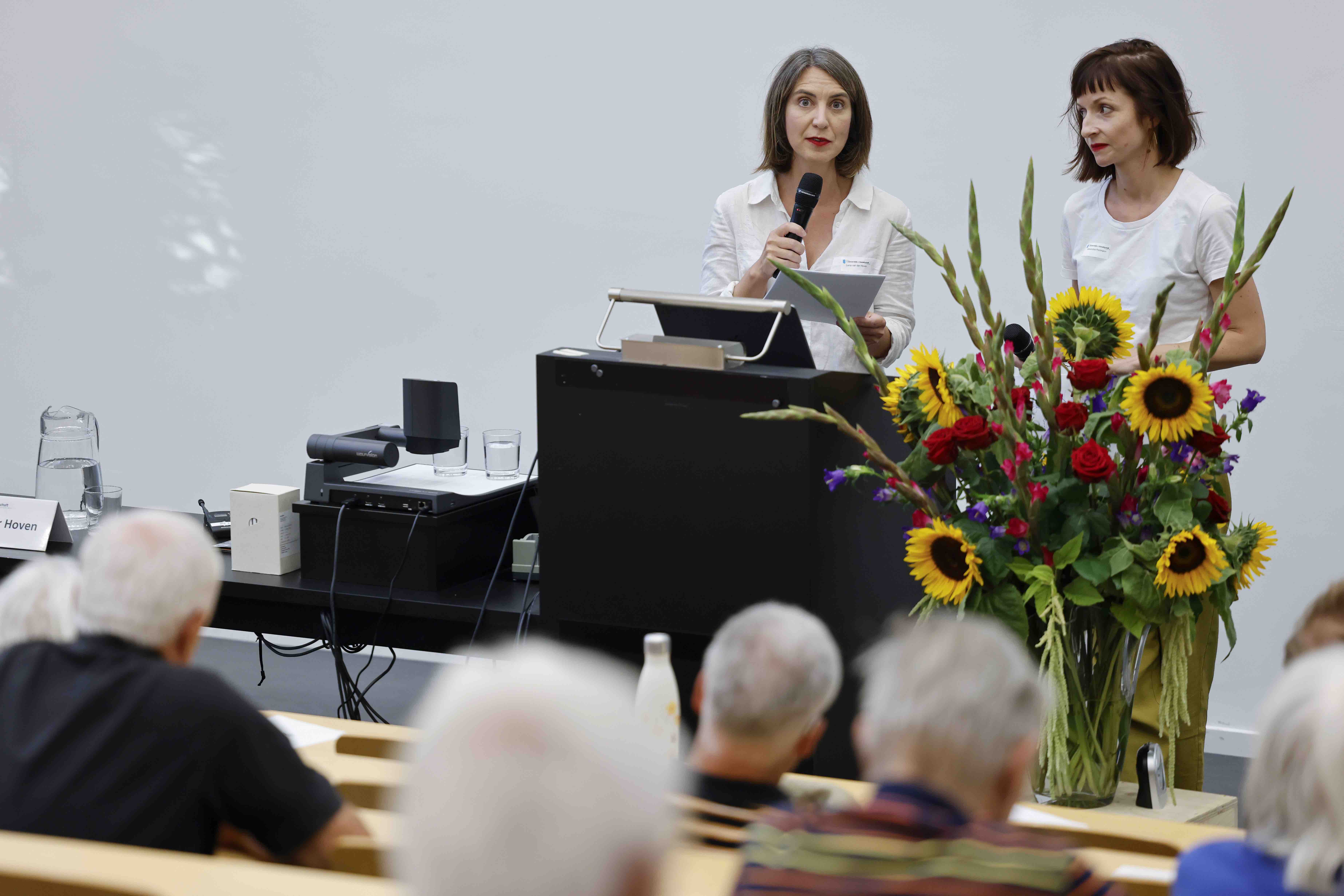Die beiden Referentinnen Alexandra Portmann und Lena van der Hosen stehen hinter einem Rednerpult. Im Vordergrund sind Zuhörer:innen sichtbar, neben dem Rednerpult steht ein Blumenstrauss.