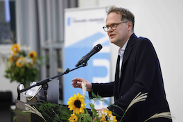 Der Referent Thomas Strässle steht hinter einem Rednerpult und spricht in ein Mikrofon. Links und rechts von ihm stehen Blumensträusse mit Sonnenblumen, im Hintergrund ist ein blauweisses Banner des Forums für Universität und Gesellschaft zu sehen.