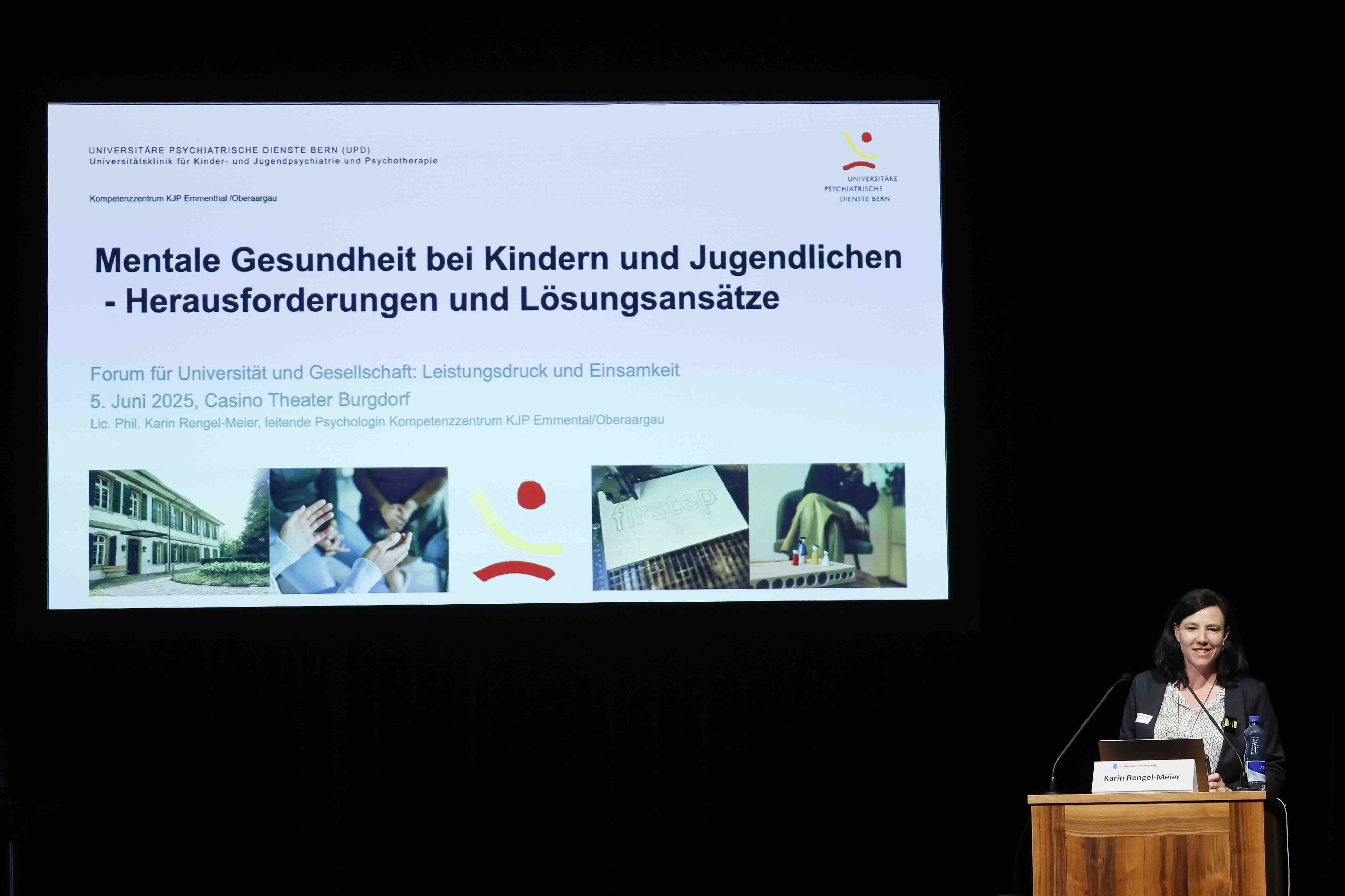 Eine Frau steht an einem Rednerpult auf einer Bühne und spricht. Hinter ihr ist eine große Leinwand zu sehen, auf der eine Präsentationsfolie projiziert wird.