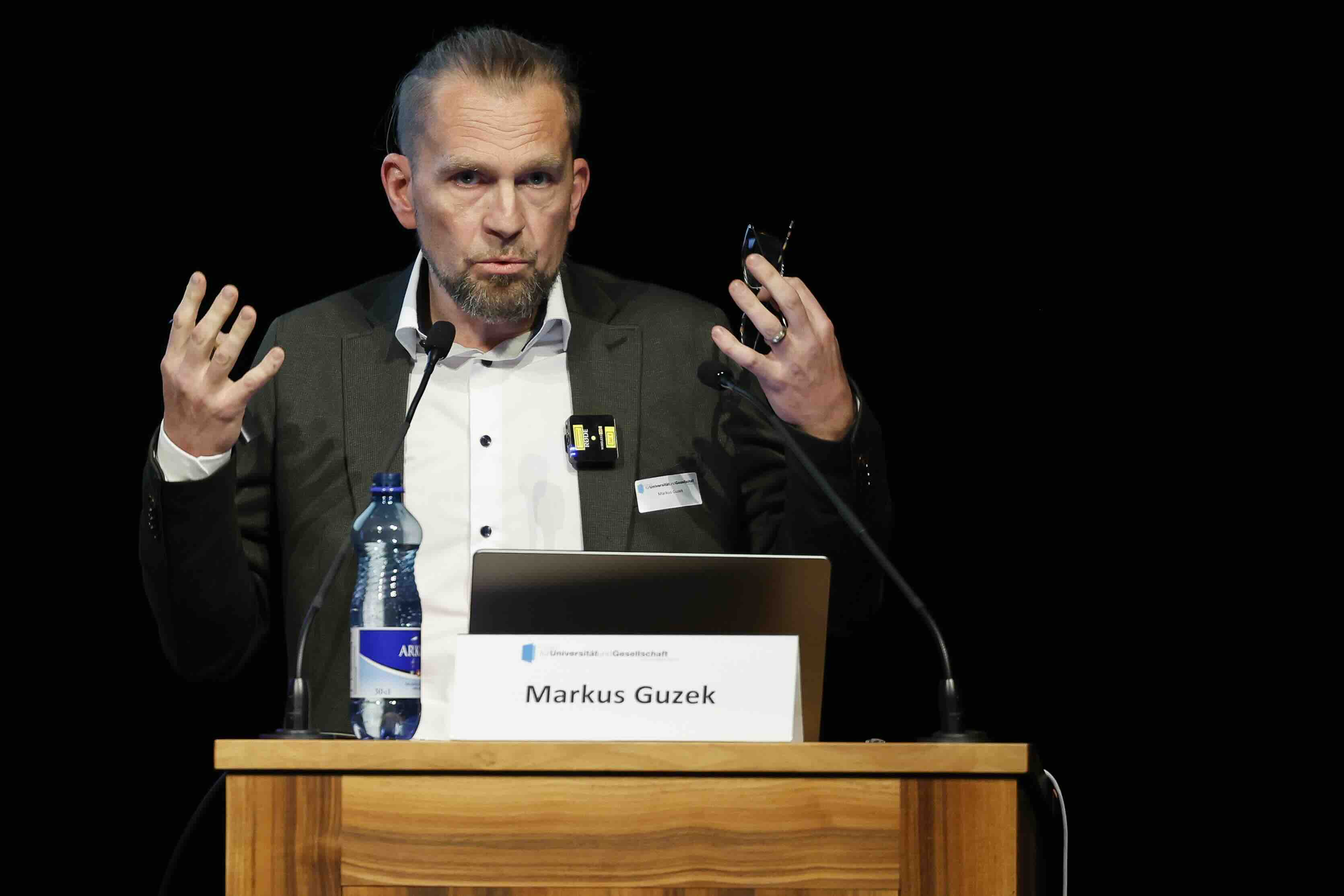 Ein Mann steht an einem Rednerpult und spricht gestikulierend mit erhobenen Händen. Vor ihm befinden sich ein Laptop und eine Wasserflasche. Auf dem Rednerpult ist ein Schild mit dem Namen „Markus Guzek“ sowie das Logo des Forums für Universität und Gesellschaft angebracht. Der Hintergrund ist dunkel, der Fokus liegt auf dem Sprecher.