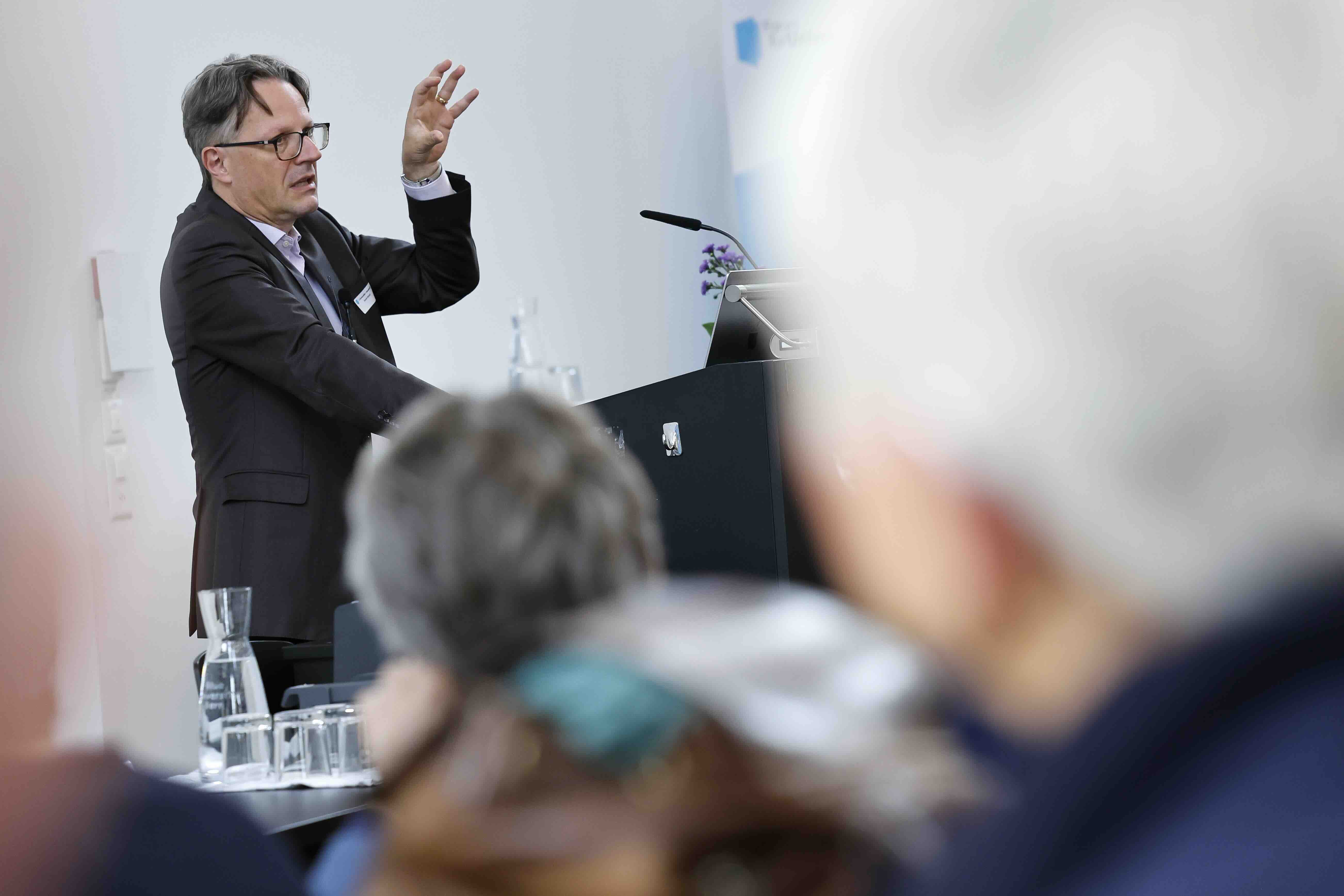 Ein Mann mit dunklem Anzug und Brille steht an einem Rednerpult und spricht zu einem Publikum. Er hebt eine Hand und gestikuliert beim Erklaeren. Im Vordergrund ist das Publikum unscharf zu sehen. Auf dem Tisch stehen Wasserkaraffen und Glaeser.