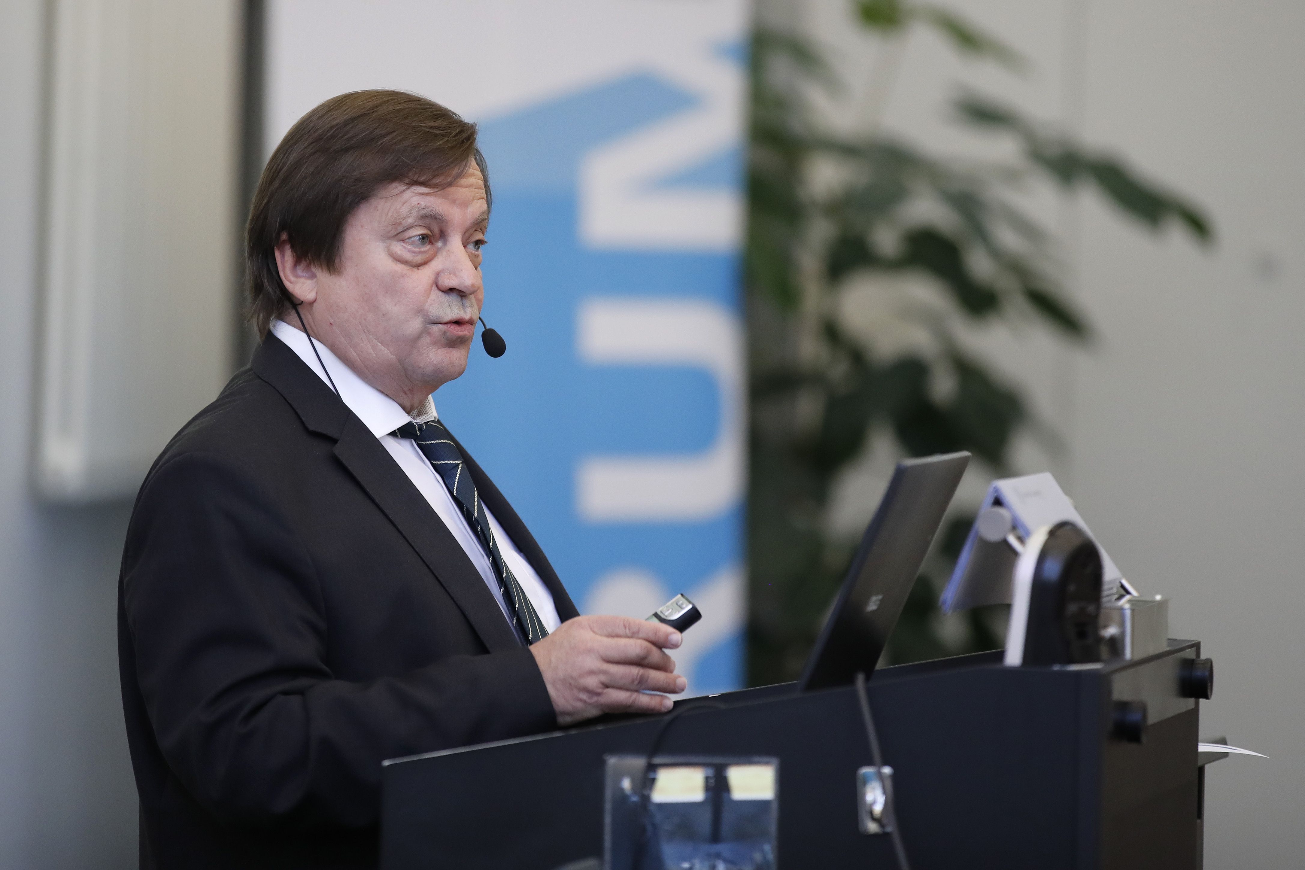 Dr. Andreas Zeller steht während seinem Referat am Rednerpult. Im Hintergrund ein blaues Plakat des Forums für Universität und Gesellschaft