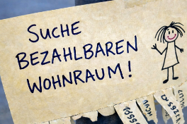 Handgeschriebener Zettel mit der Aufschrift "Suche bezahlbaren Wohnraum!" neben einer einfachen Kinderzeichnung einer Person; im Hintergrund sind unscharfe Wohnungsanzeigen zu sehen. Das Bild symbolisiert die dringende Suche nach günstigem, bezahlbaren Wohnraum und thematisiert die aktuelle Wohnungsnot.
