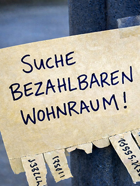 Das Bild zeigt ein handgeschriebenes Inserat mit dem Text „Suche bezahlbaren Wohnraum!“ an einer Pinnwand oder Litfasssäule. Unten befinden sich mehrere abreissbare Kontaktstreifen. Das Motiv steht symbolisch für Wohnungsknappheit, hohe Mietpreise und die angespannte Lage auf dem Wohnungsmarkt.