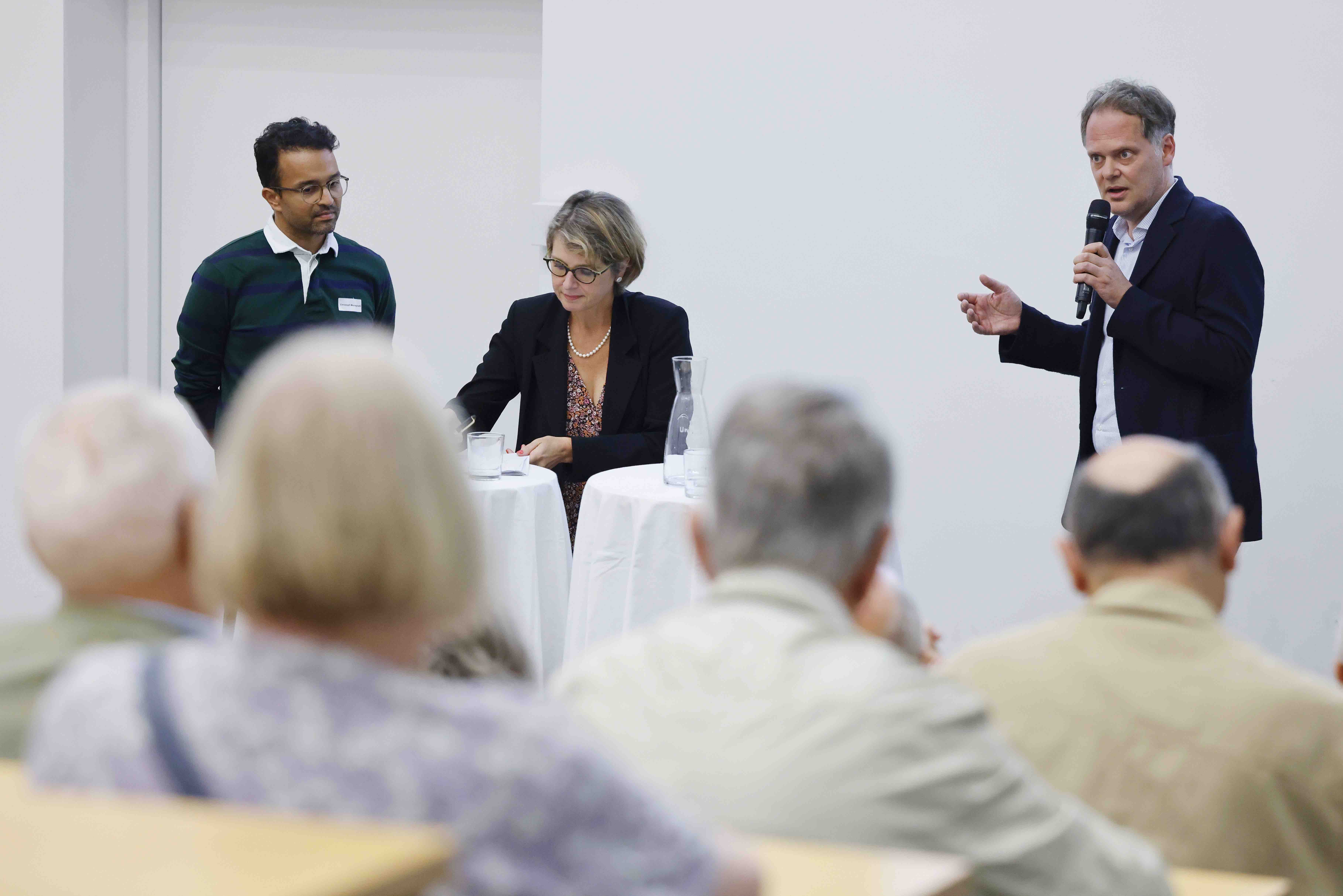 Drei Personen stehen vor dem Publikum und beantworten Fragen. Der Mann rechts aussen spricht in ein Handmikrofon, die Frau in der Mitte notiert sich etwas. Im Vordergrund ist ein Teil des Publikums von hinten sichtbar.