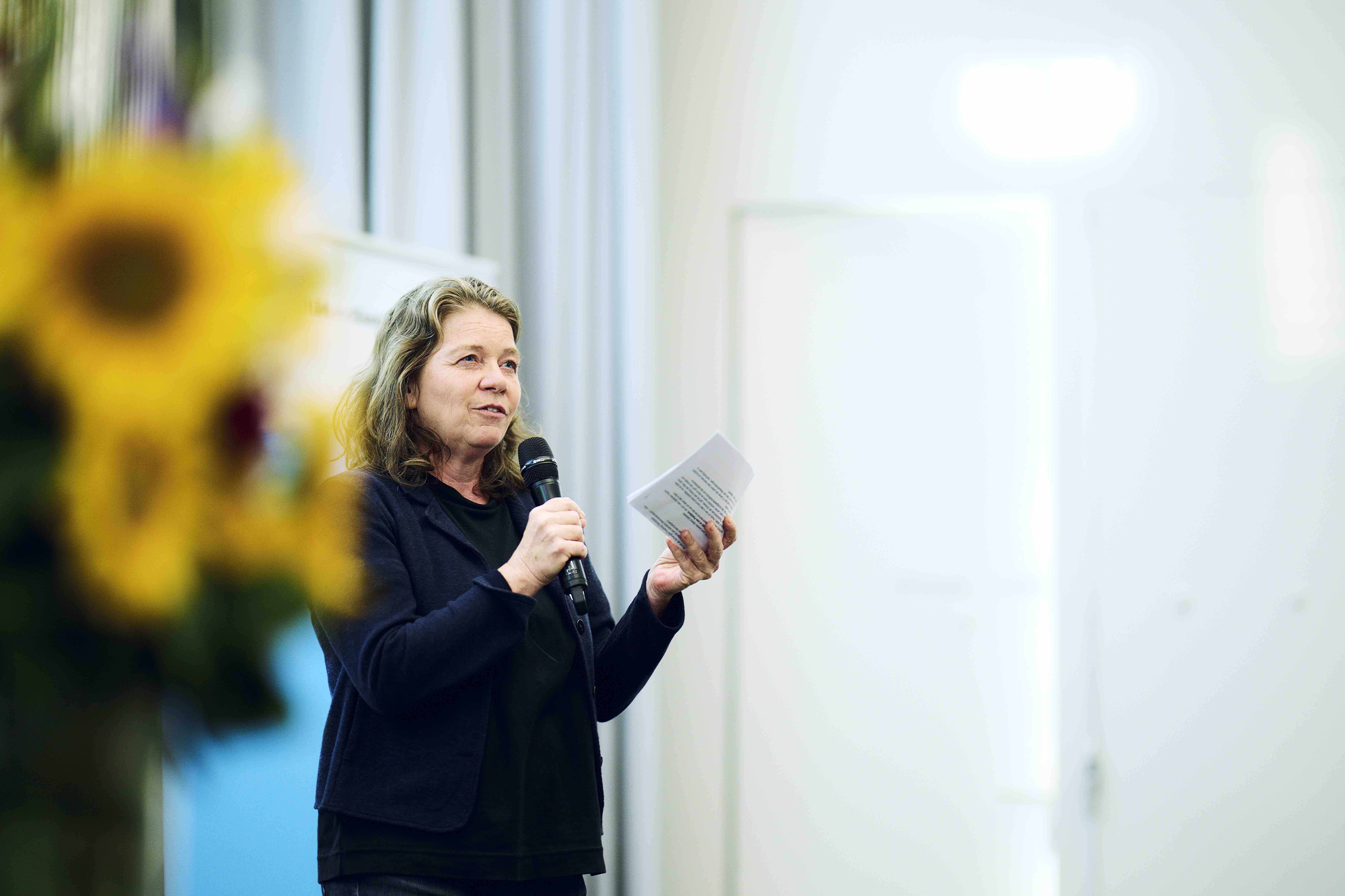 Die Moderatorin Dagmar Walser hält in der einen Hand ihr Manuskript und in der anderen ein Mikrofon. Sie ist schwarz gekleidet, links neben ihr ist umscharf ein Strauss Sonnenblumen erkennbar.