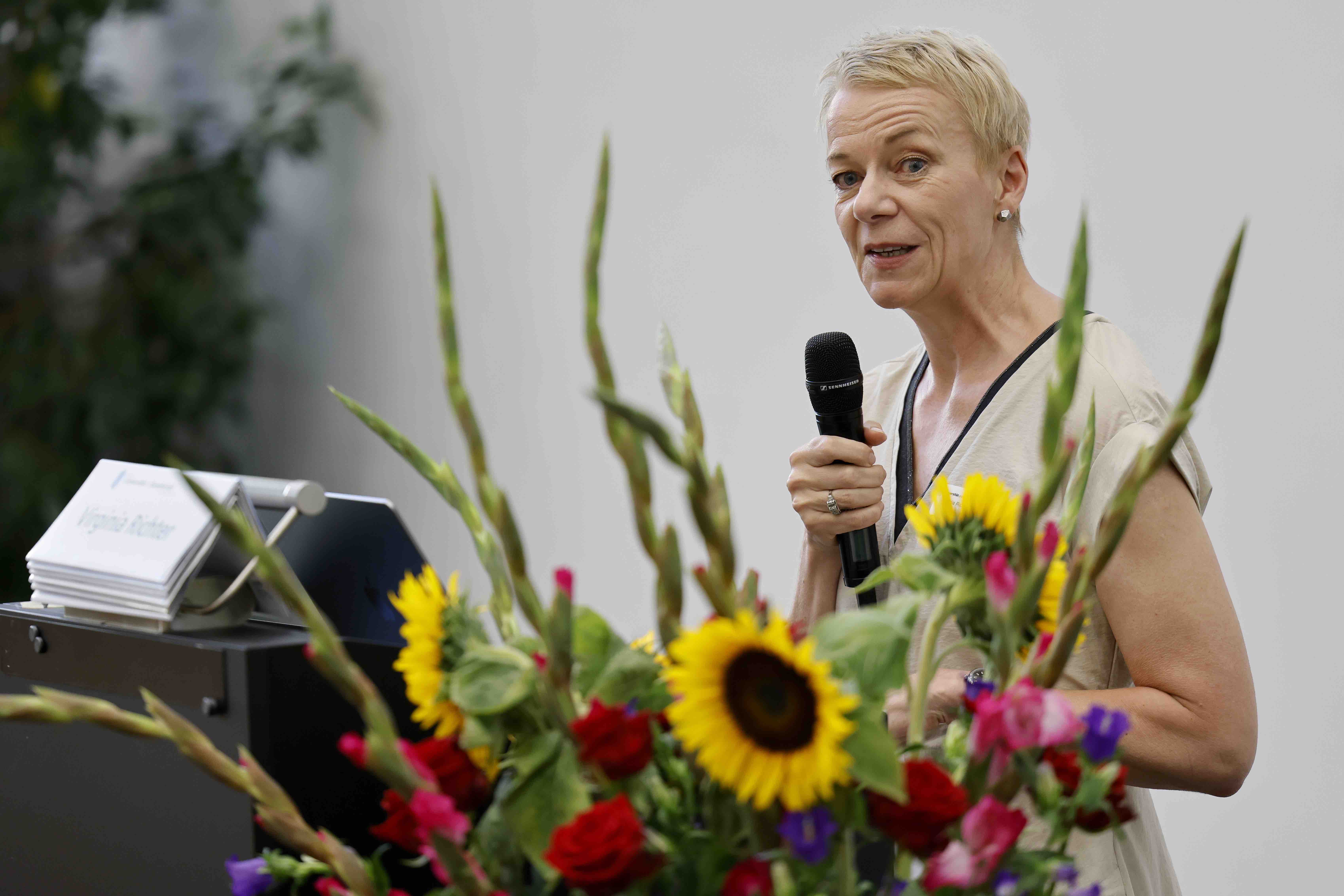 Forumspräsidentin Prof. Dr. Virginia Richter steht an einem Rednerpult und hält ein Mikrofon in der linken Hand. Im Vordergrund steht ein Blumenstrauss mit Sonnenblumen und pinken Gladiolen.