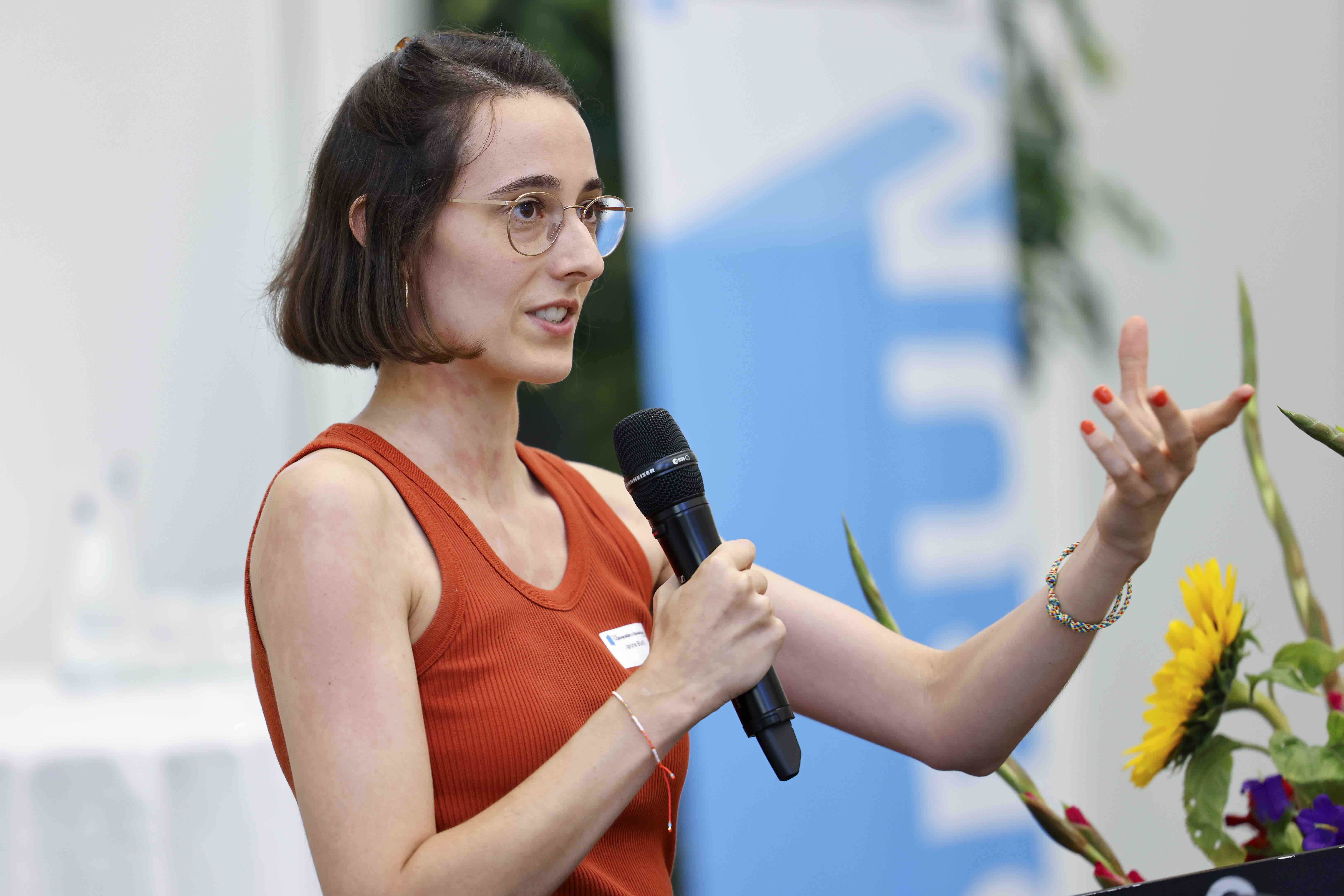 Die Referentin Janine Bürkli spricht in ein Handmikrofon und gestikuliert mit der linken Hand. Im Hintergrund ist das blauweisse Banner mit dem Forumslogo erkennbar.
