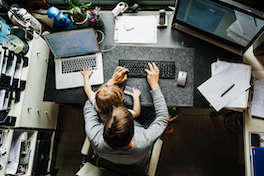 Die Arbeitswelt im Wandel: Sicht von oben auf einen Menschen, der mit Kind im Homeoffice an einem Schreibtisch sitzt und am Computer arbeitet.