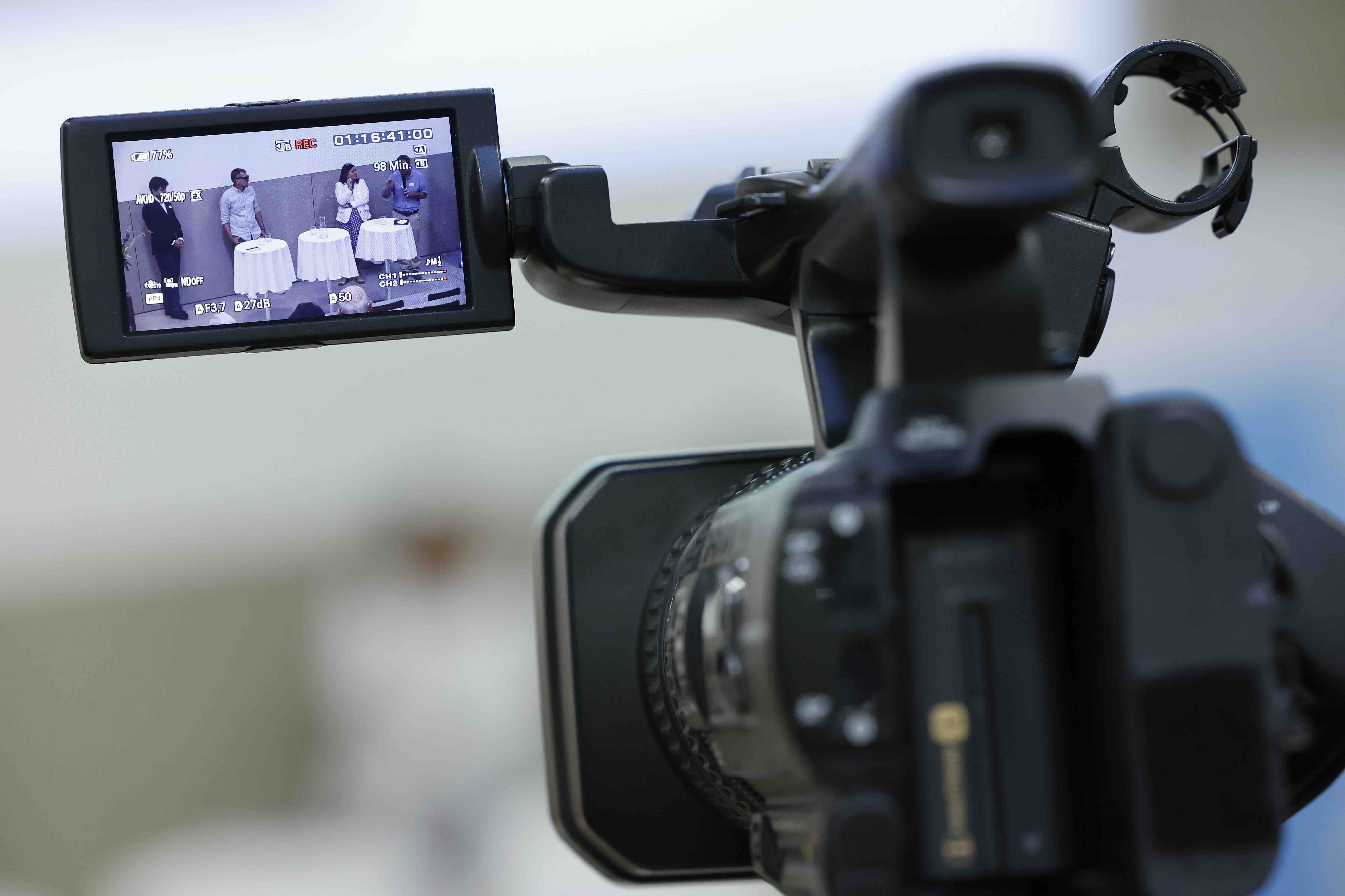 Aufnahme des Displays einer Filmkamera. Auf dem Display sind vier Personen zu sehen, die während einer Podiumsdiskussion zum Thema Arbeitswelt im Wandel hinter drei Stehtischen mit weissen Tischtüchern stehen.