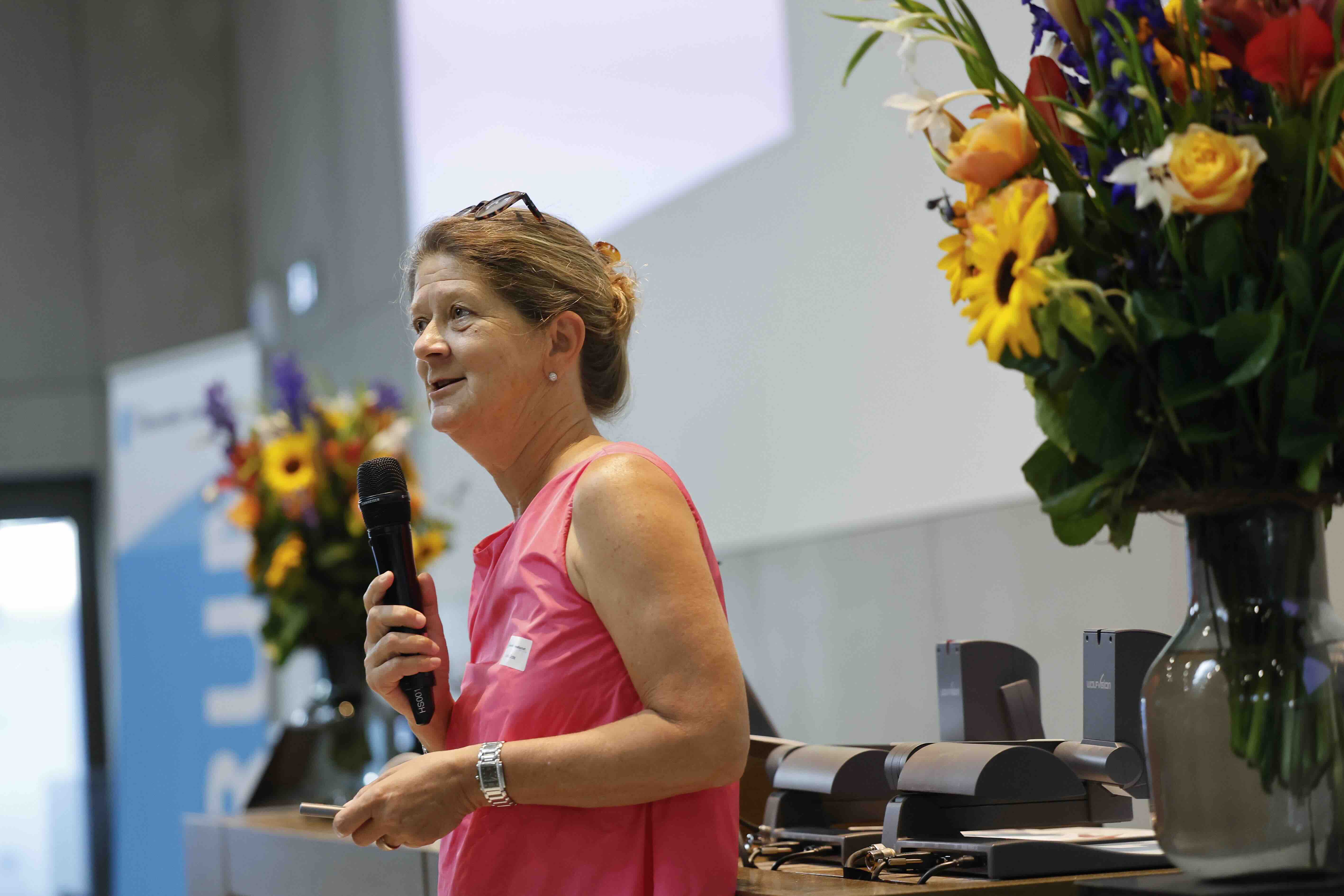 Prof. Dr. Gudela Grote spricht in ein Handmikrofon, in der linken Hand hält sie einen Presenter. Sie trägt ein pinkfarbenes Kleid, rechts und links von ihr steht je ein Blumenstrauss.