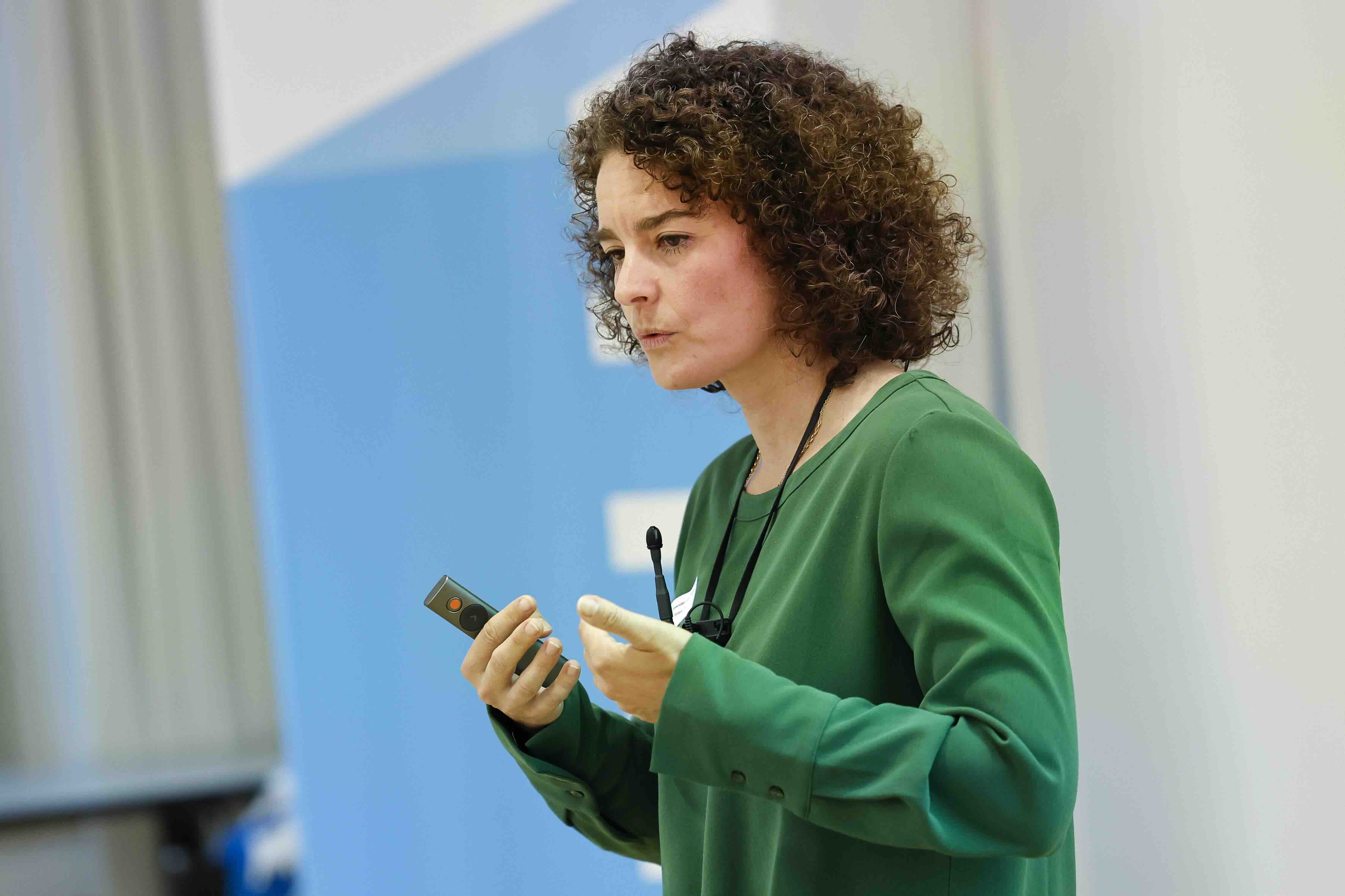 Eine Frau mit lockigen Haaren hält einen Vortrag über Beratungsangebote zur mentalen Gesundheit und spricht mit einem Headset-Mikrofon, während sie eine Präsentationsfernbedienung in der Hand hält.