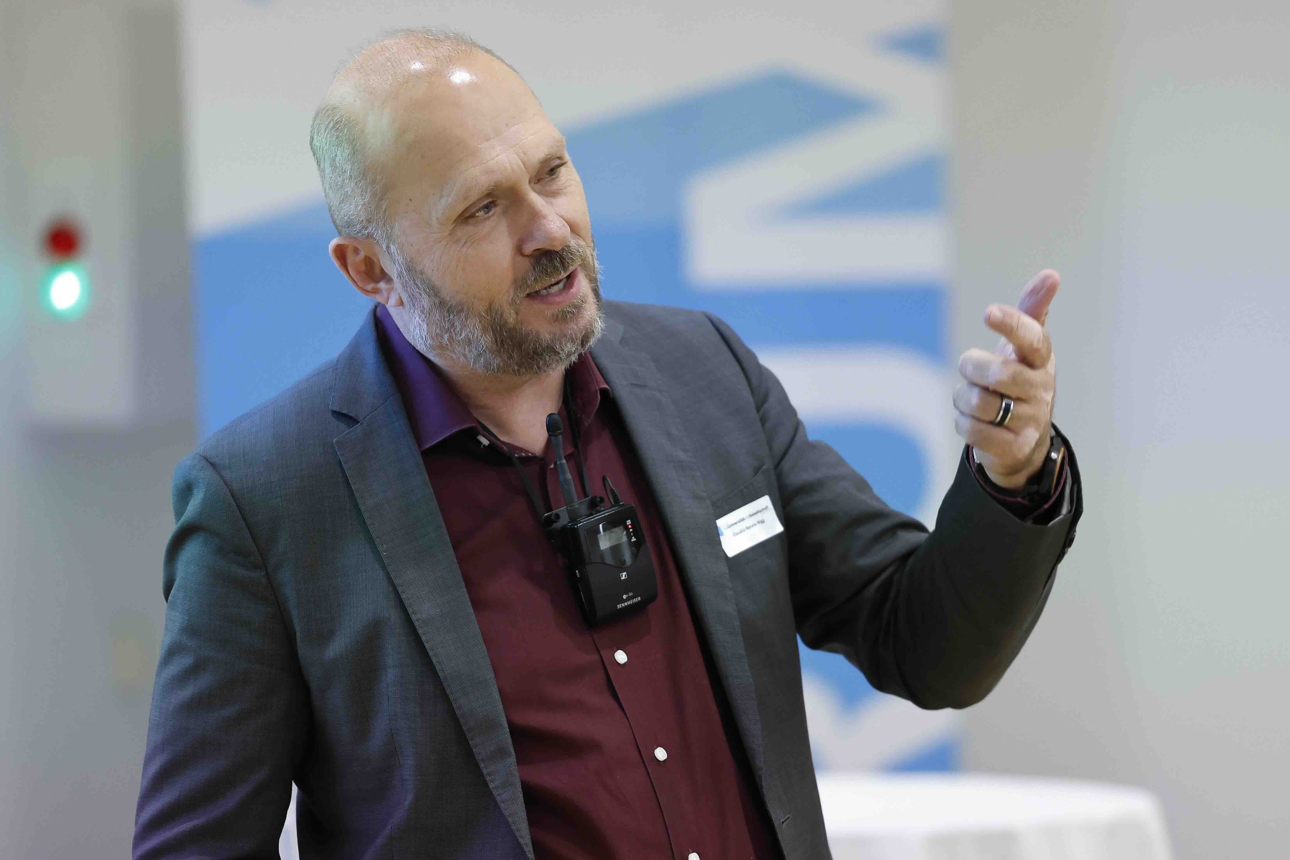Ein Mann mittleren Alters hält während eines Vortrags eine Rede. Er trägt ein Headset-Mikrofon, ein weinrotes Hemd und ein graues Sakko. Mit einer Handgeste unterstreicht er seine Worte.