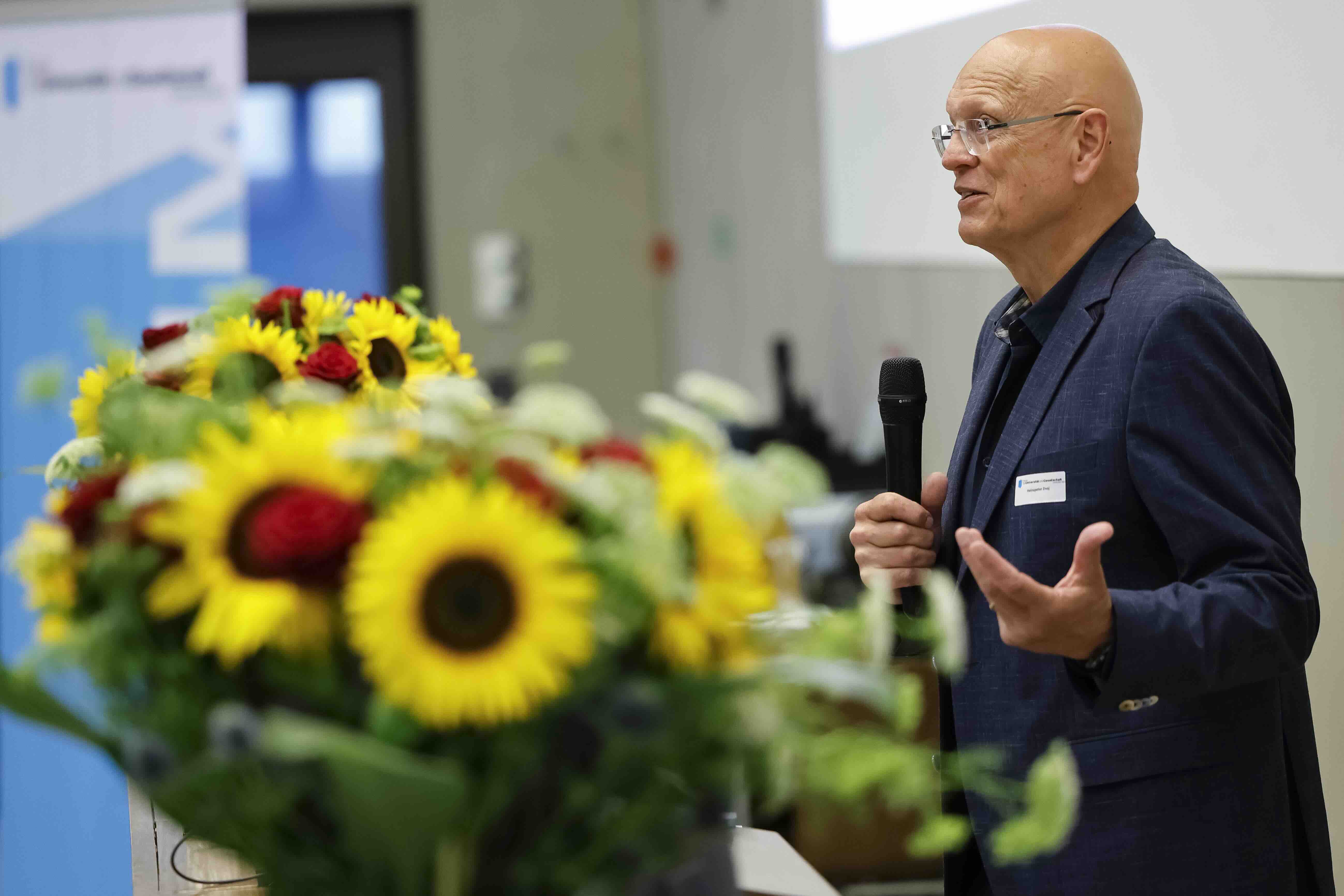 Heinzpeter Znoj spricht mit Mikrofon am Rednerpult vor einem Blumenschmuck aus Sonnenblumen.