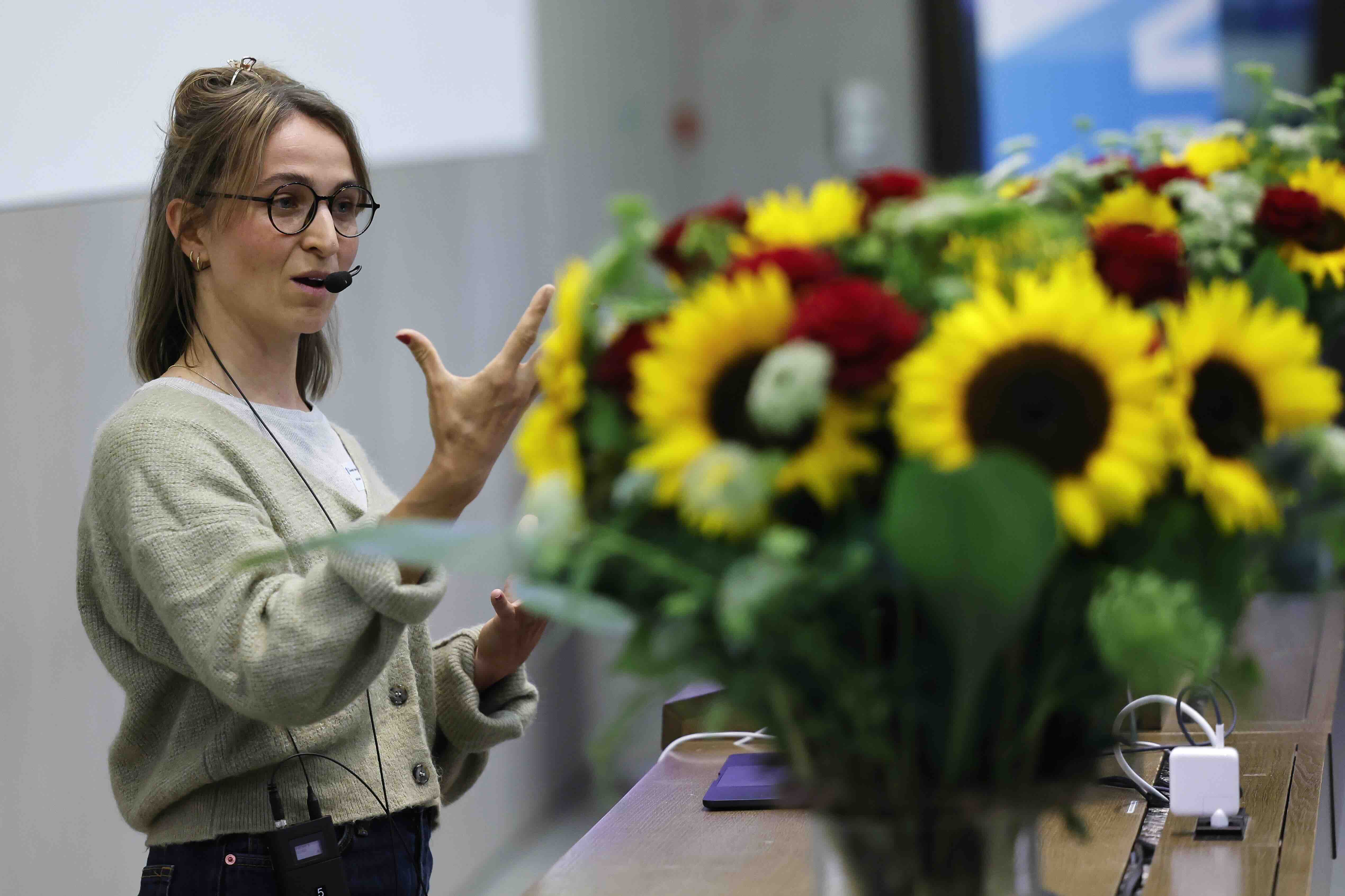 Noemi Seewer spricht mit Headset-Mikrofon gestikulierend über Einsamkeit, vor einem Pult mit Sonnenblumen.