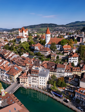 Luftaufnahme der Thuner Altstadt mit Aare, Schloss Thun, Stadtkirche und dicht bebautem Stadtzentrum vor der hügeligen Landschaft des Berner Oberlands.