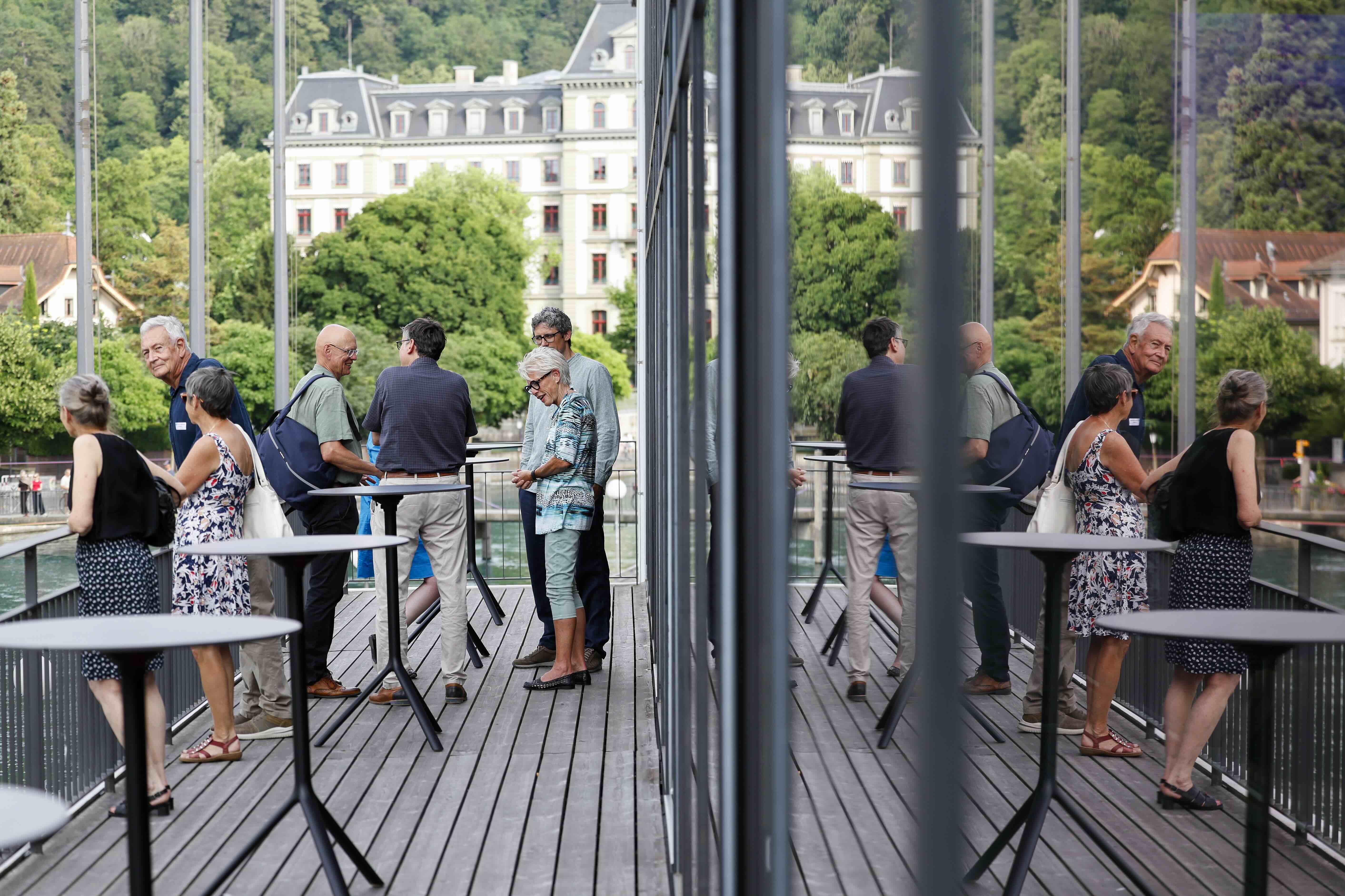 Gäste tauschen sich auf der Terrasse des Hotel Aare Thun mit Blick auf die Aare aus. Im Hintergrund sind viele Bäume und ein grosses Haus zu sehen.