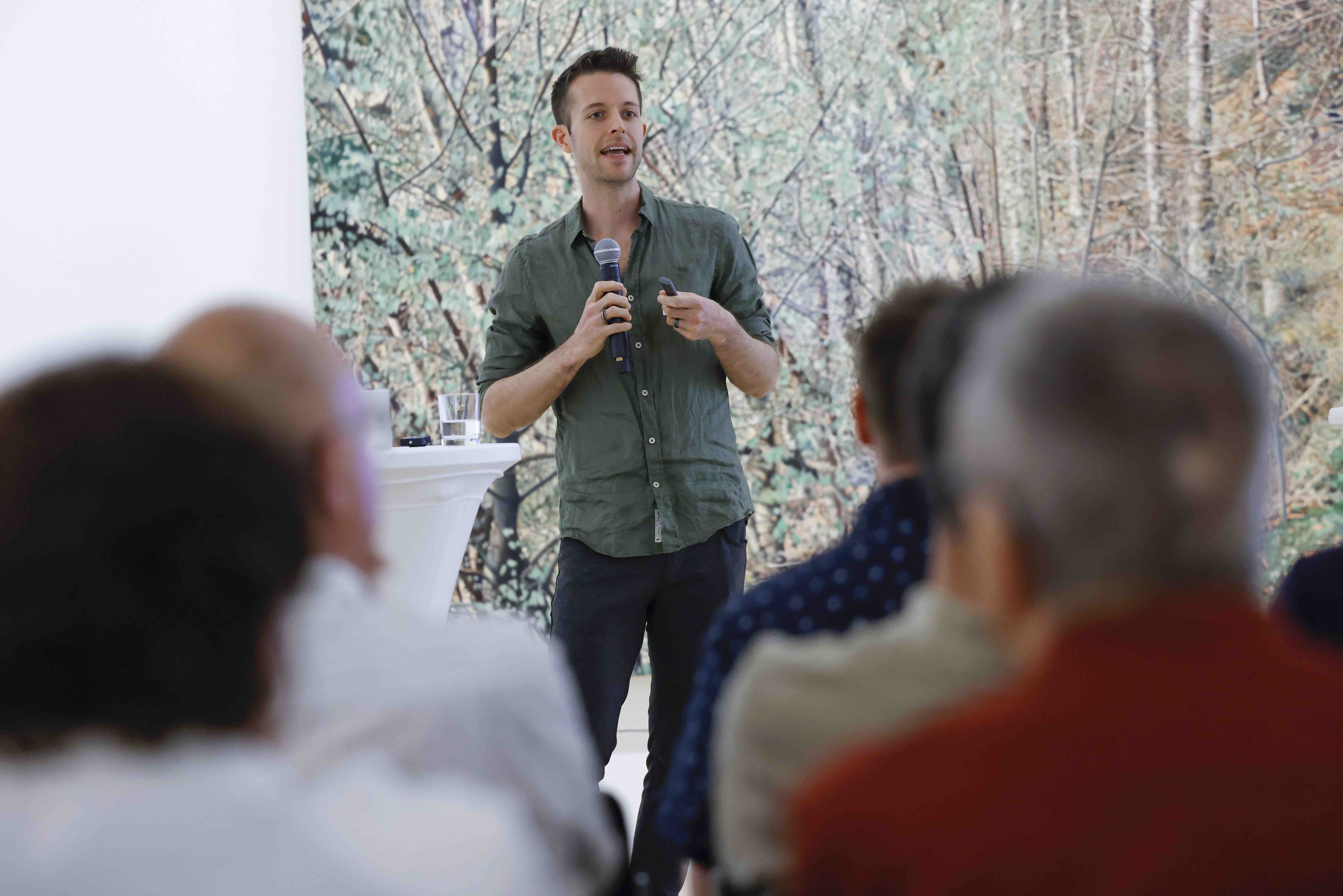 Blick durch die Publikumsreihen auf den Referenten Tobias Rohrbach. Er steht vor dem Bild «Frühling» von Franz Gertsch.