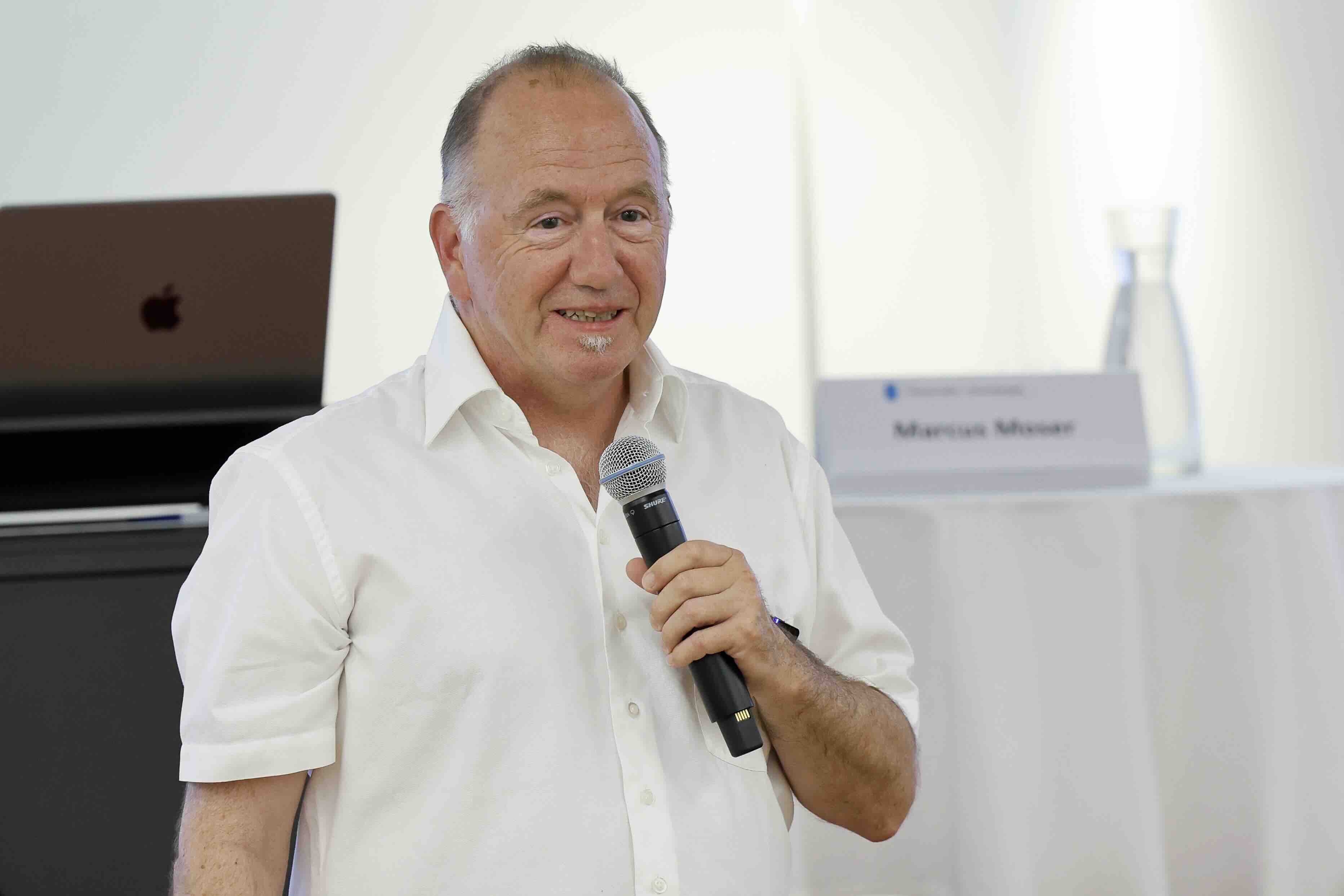 Ein Mann in weissem Hemd steht vor einem Laptop und einem Rednerpult. Er hält ein Mikrofon und spricht lächelnd in einem hellen Raum