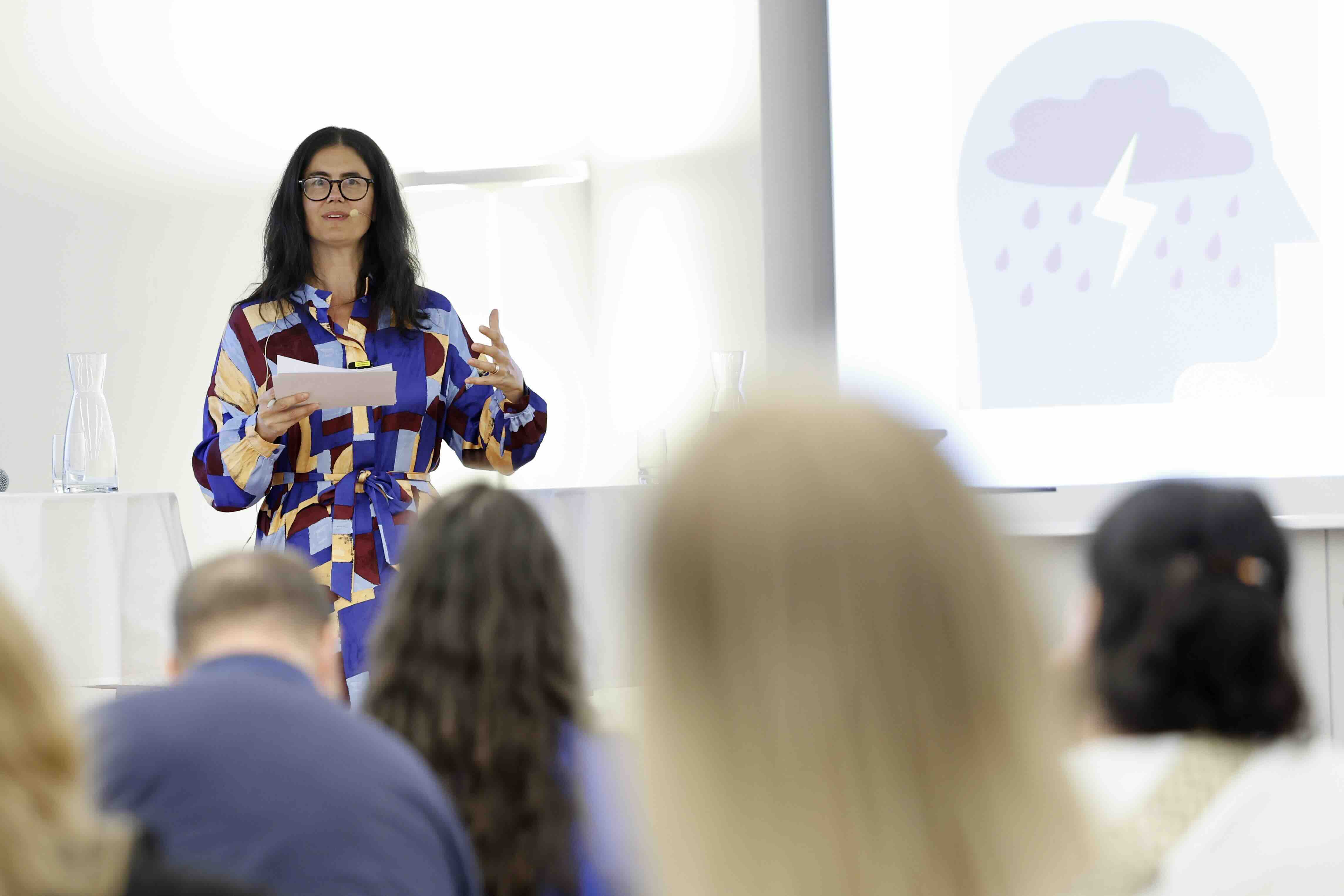 Eine Frau mit langen dunklen Haaren und bunter Bluse spricht vor Publikum. Sie hält Moderationskarten in der Hand und steht vor einer Projektion mit einer Grafik eines Kopfes mit Wolke, Blitz und Regentropfen.