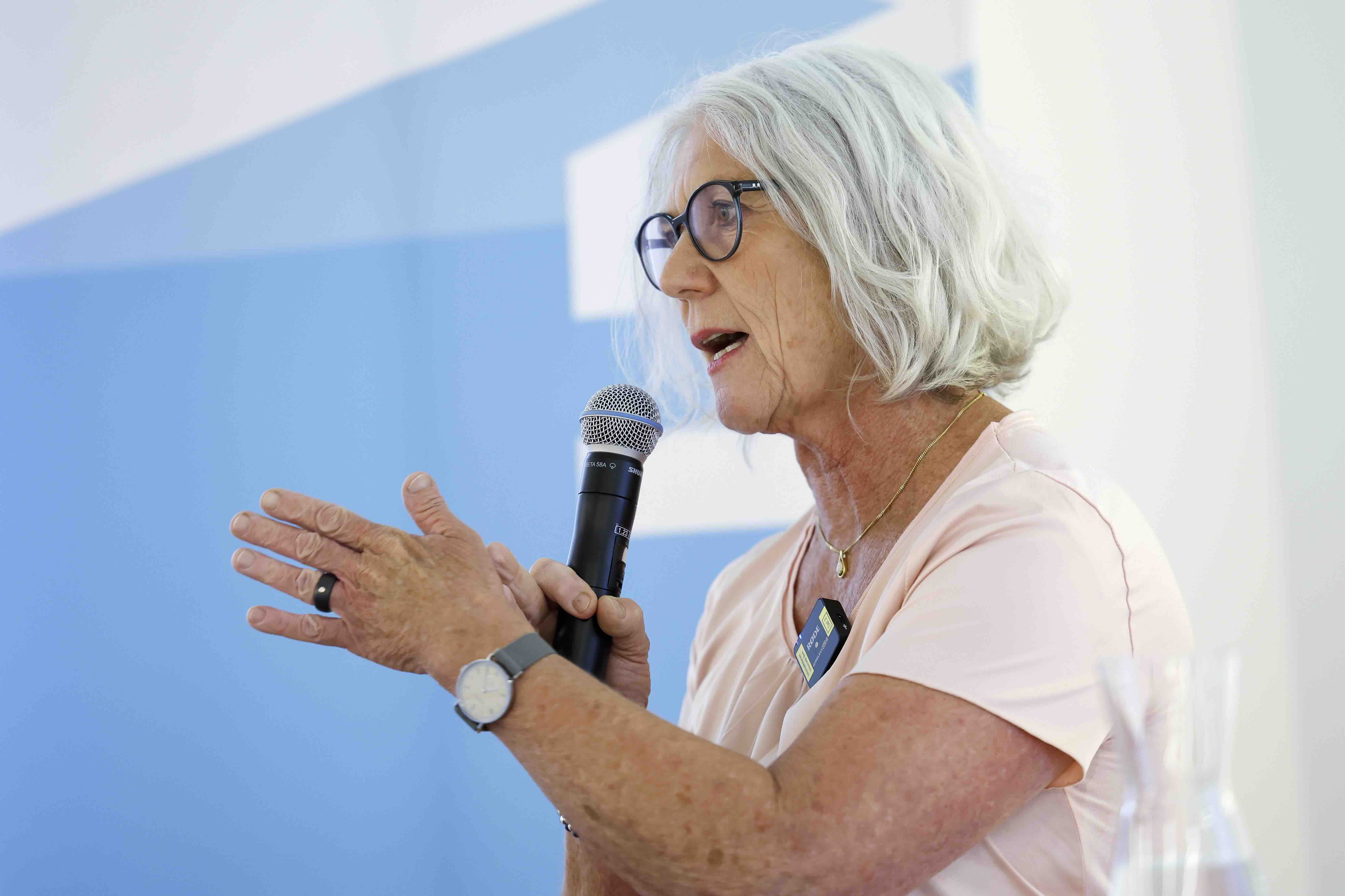 Eine ältere Frau mit grauen Haaren und Brille spricht in ein Mikrofon. Sie trägt ein helles Oberteil, eine Armbanduhr und einen Ring. Im Hintergrund ist eine blaue Wand mit geometrischem Muster zu sehen.