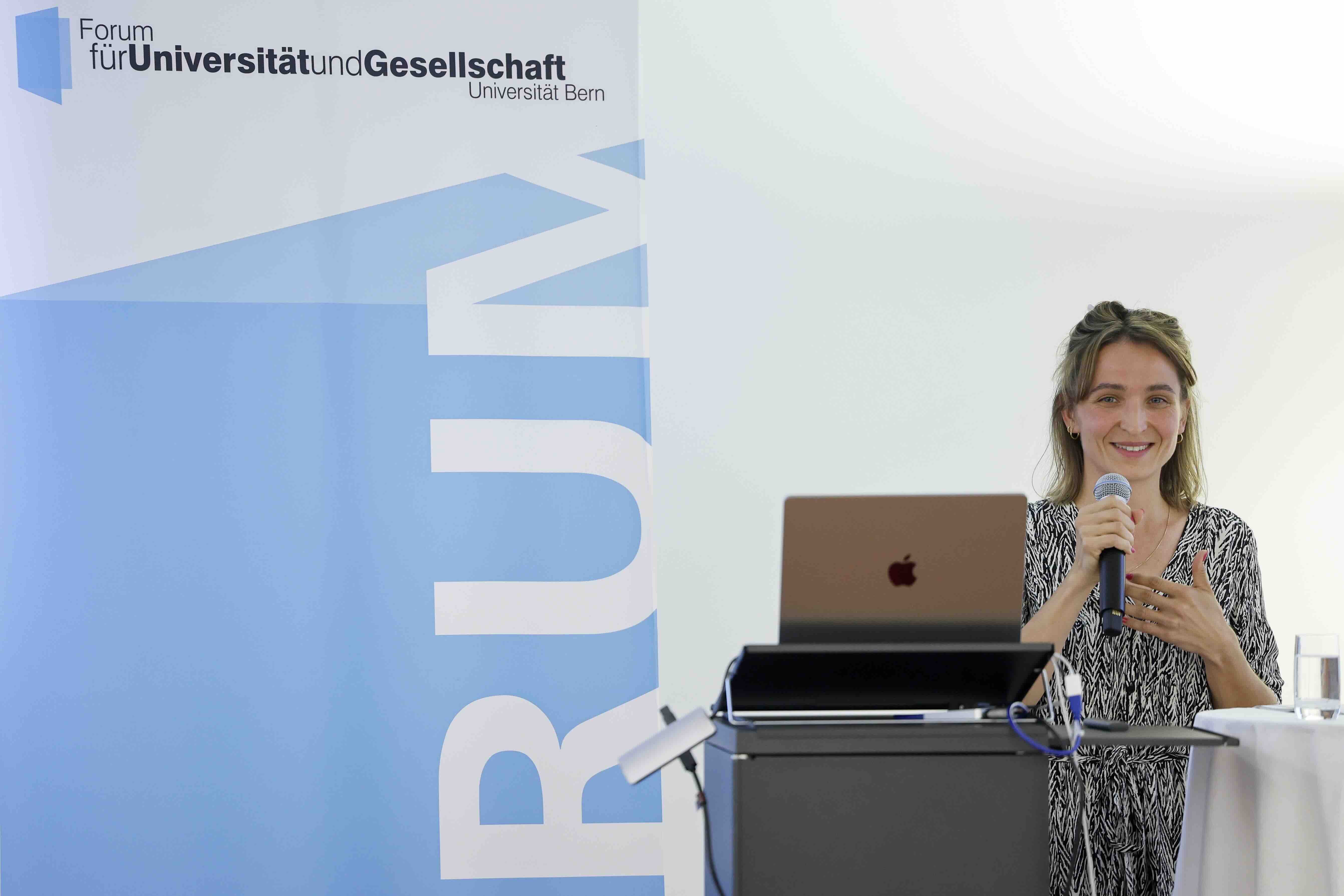 Eine junge Frau mit langen, offenen Haaren spricht lächelnd in ein Mikrofon. Sie steht hinter einem Rednerpult mit Laptop und einem Glas Wasser. Links im Bild ist ein Roll-Up mit dem Logo des Forums für Universität und Gesellschaft der Universität Bern zu sehen.