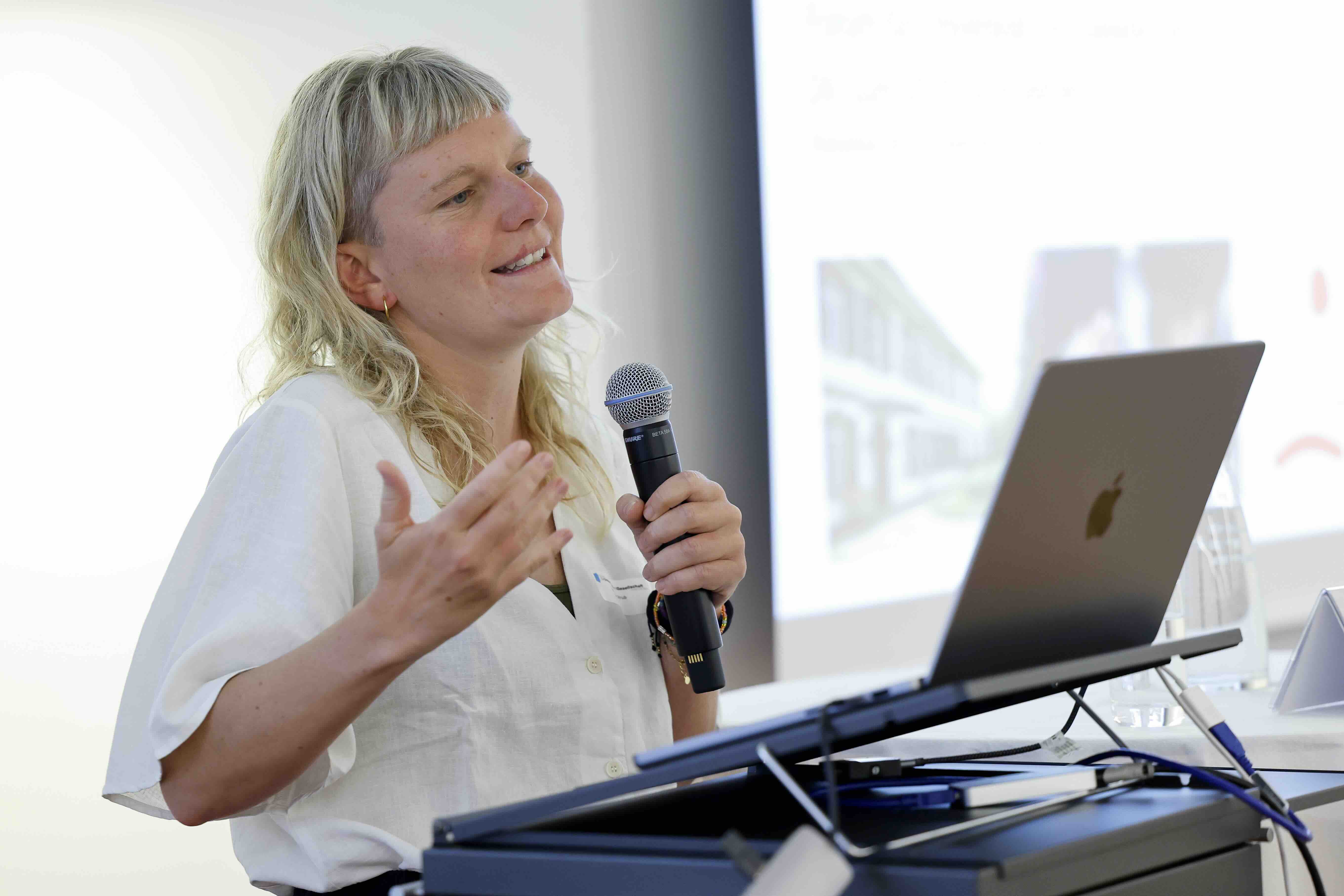Eine Frau mit mittellangem, blondem Haar und hellem Hemd spricht mit einem Mikrofon. Sie steht hinter einem Laptop auf einem Rednerpult und gestikuliert mit der freien Hand. Im Hintergrund ist eine Präsentationsfolie zu sehen.