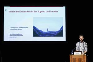 Ein junger Mann im gestreiften Hemd steht am Rednerpult. Auf der Leinwand hinter ihm ist eine Präsentationsfolie mit dem Titel „Wider die Einsamkeit in der Jugend und im Alter“ und einer Illustration einer allein stehenden Person in einer abstrakten Landschaft zu sehen.