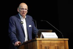 Ein älterer Mann mit Glatze und Brille spricht am Rednerpult. Er trägt ein Mikrofon am Revers und blickt in Richtung Publikum. Vor ihm steht ein Laptop und ein Schild mit seinem Namen.