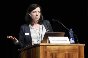 Eine Frau mit dunklem Haar spricht am Rednerpult und gestikuliert mit der linken Hand. Vor ihr stehen ein Laptop und ein Namensschild, auf dem ihr Name zu lesen ist.