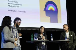 Vier Personen stehen und sitzen an zwei Stehtischen auf der Bühne. Eine Frau moderiert, während die anderen drei diskutieren. Im Hintergrund sind die Namen der Diskutierenden und ein farbiges Kopf-Symbol eingeblendet.