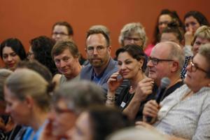 Eine Frau mit Mikrofon spricht aus dem Publikum. Neben ihm sitzen mehrere aufmerksam zuhörende Personen. Im Vordergrund ist das Publikum unscharf zu sehen.