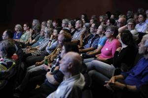 Das Publikum sitzt dicht beieinander in mehreren Reihen und hört aufmerksam zu. Die meisten Personen tragen Namensschilder, die Stimmung ist konzentriert.