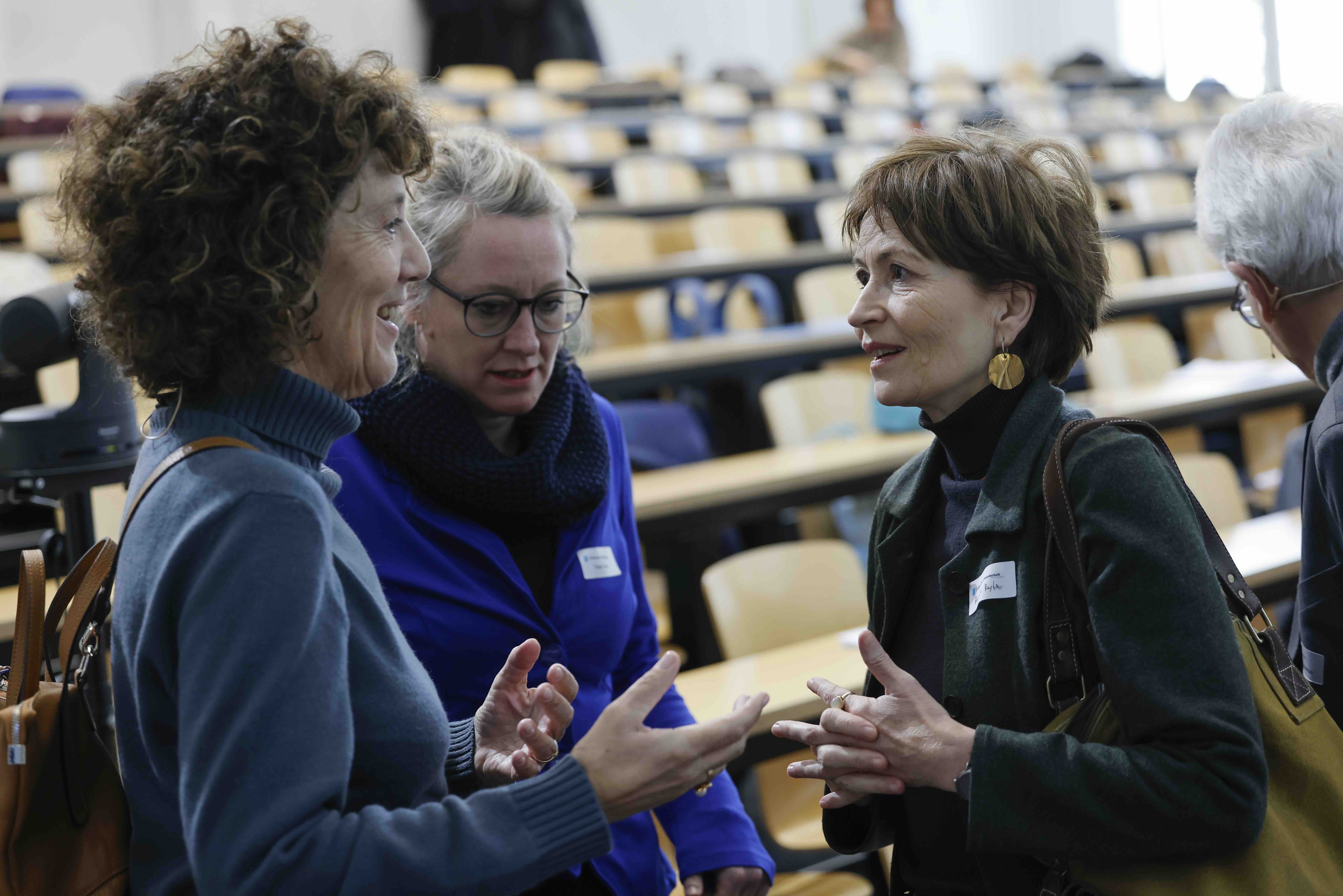 Drei Frauen stehen vor leeren Hörsaalreihen und sind in eine Diskussion vertieft.