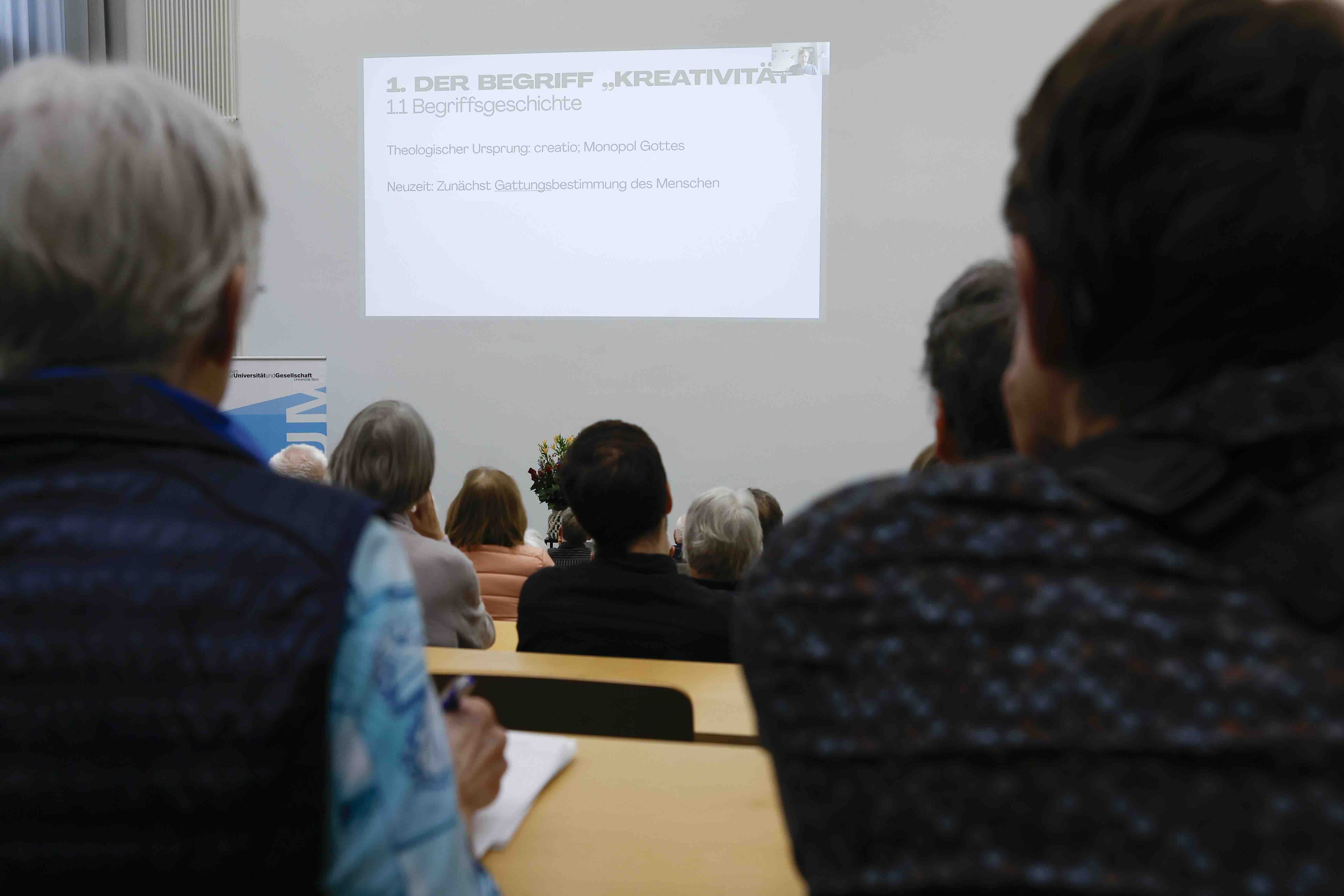 Sicht von hinten auf Menschen, die in einem Hörsaal sitzen. An die Wand vor ihnen wird eine Präsentation projiziert.